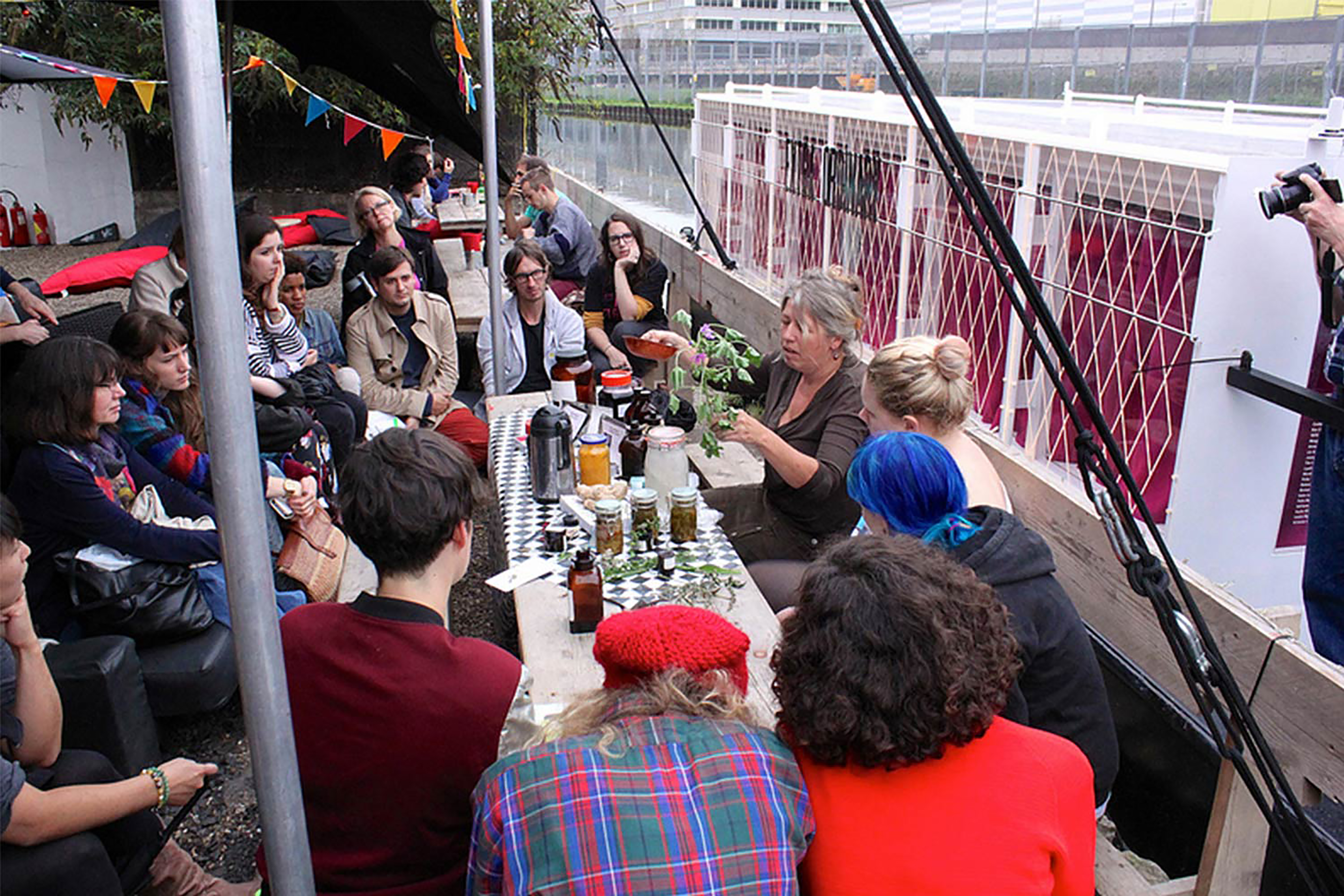 A large group of people sitting on a bank under a large canopy watching a demonstration being led by two women, next to a large white canal boat.