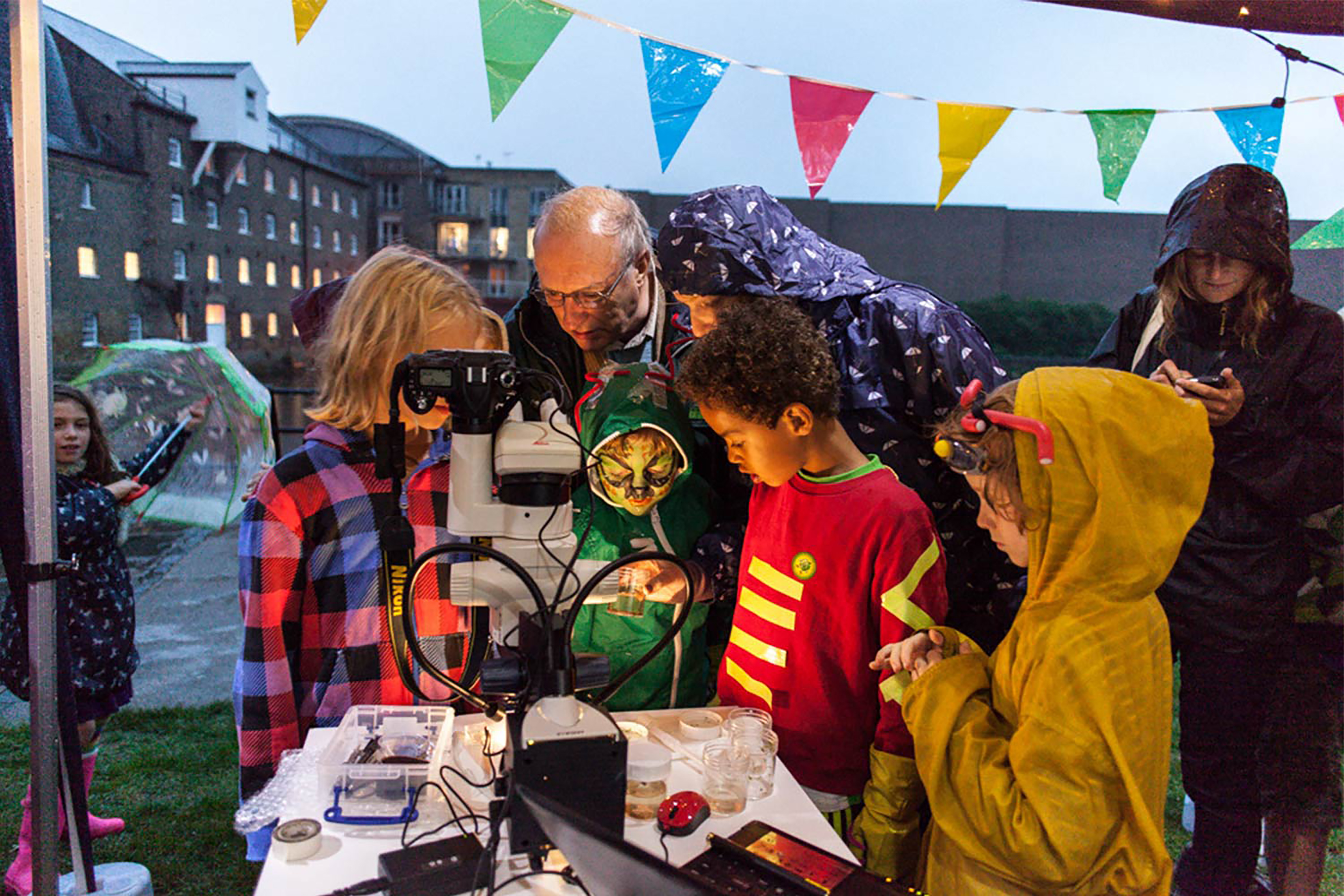 Two adults and three children leaning down looking over a table containing a large grey camera, under a canopy with colourful festival banners.