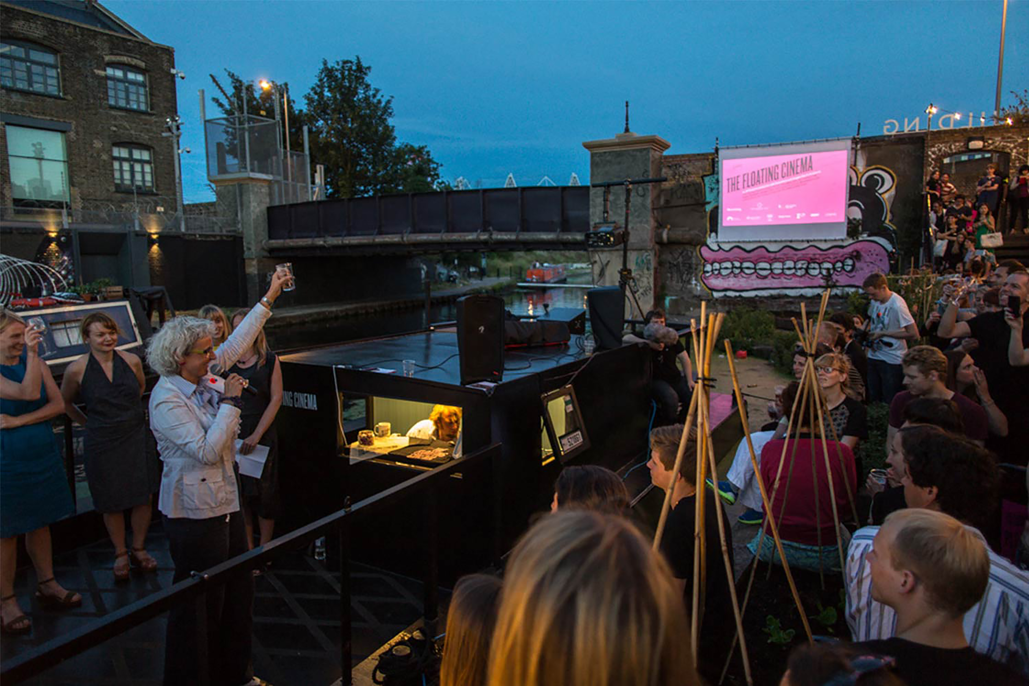A large barge canal on a bank next to a large group of people at night with a large projection screen in the background showing a film.