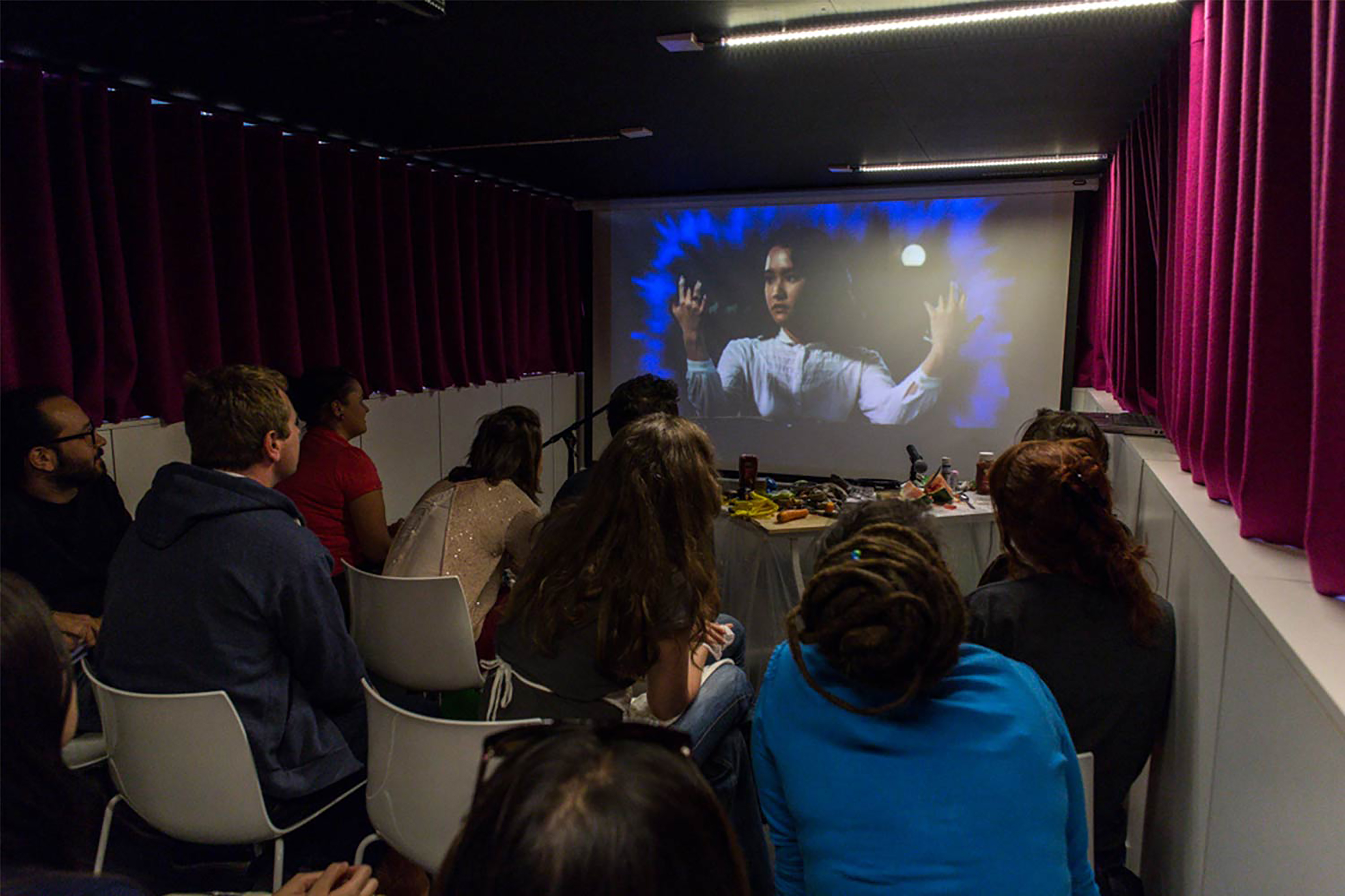 A group of people in a small room with purple curtains all watching a small projection screen showing a film.