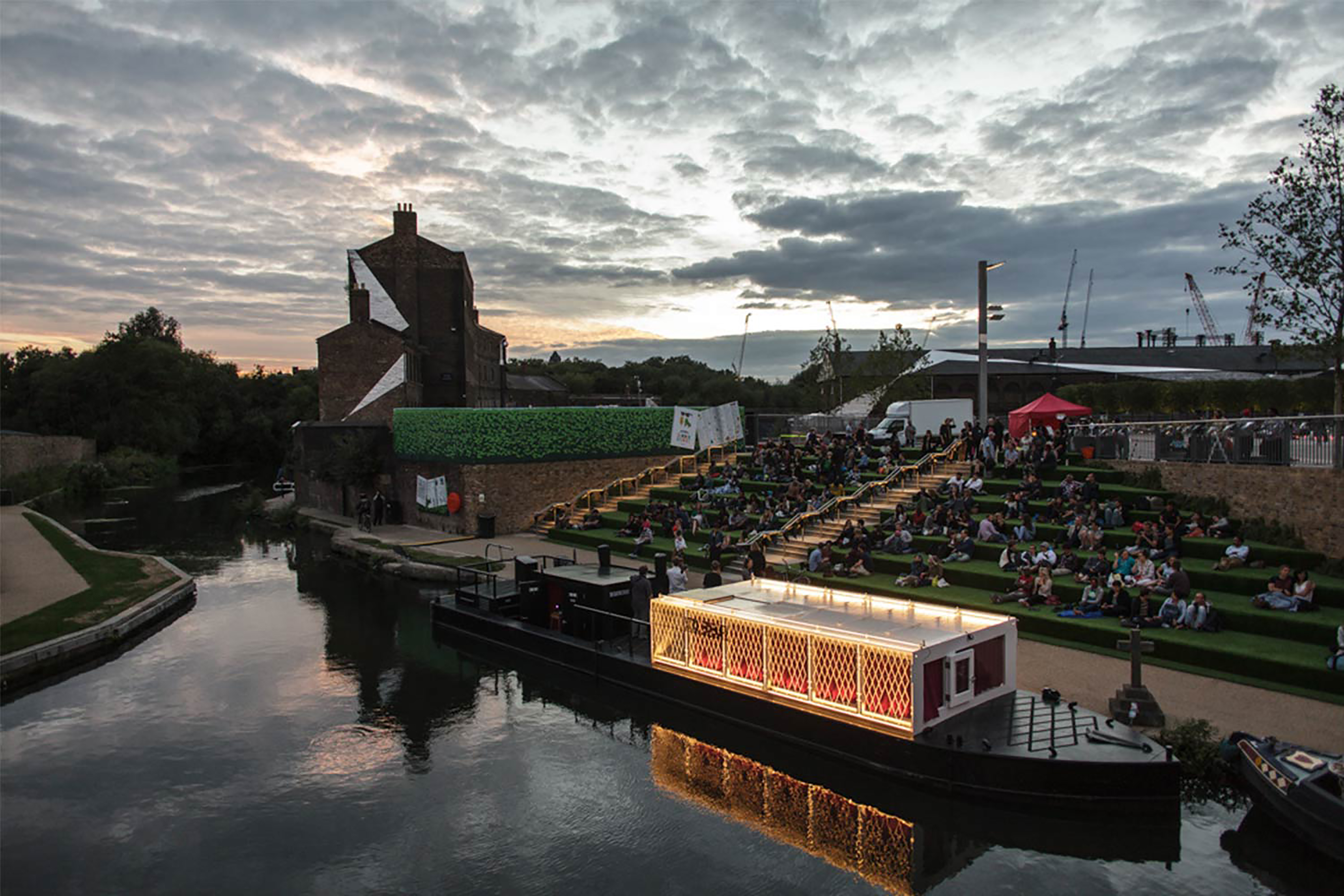 A long grey boat with a wooden boxed top parked up next to a bank of steps, with green grass, a large group of people sitting around, in front of a large red brick building at dusk.