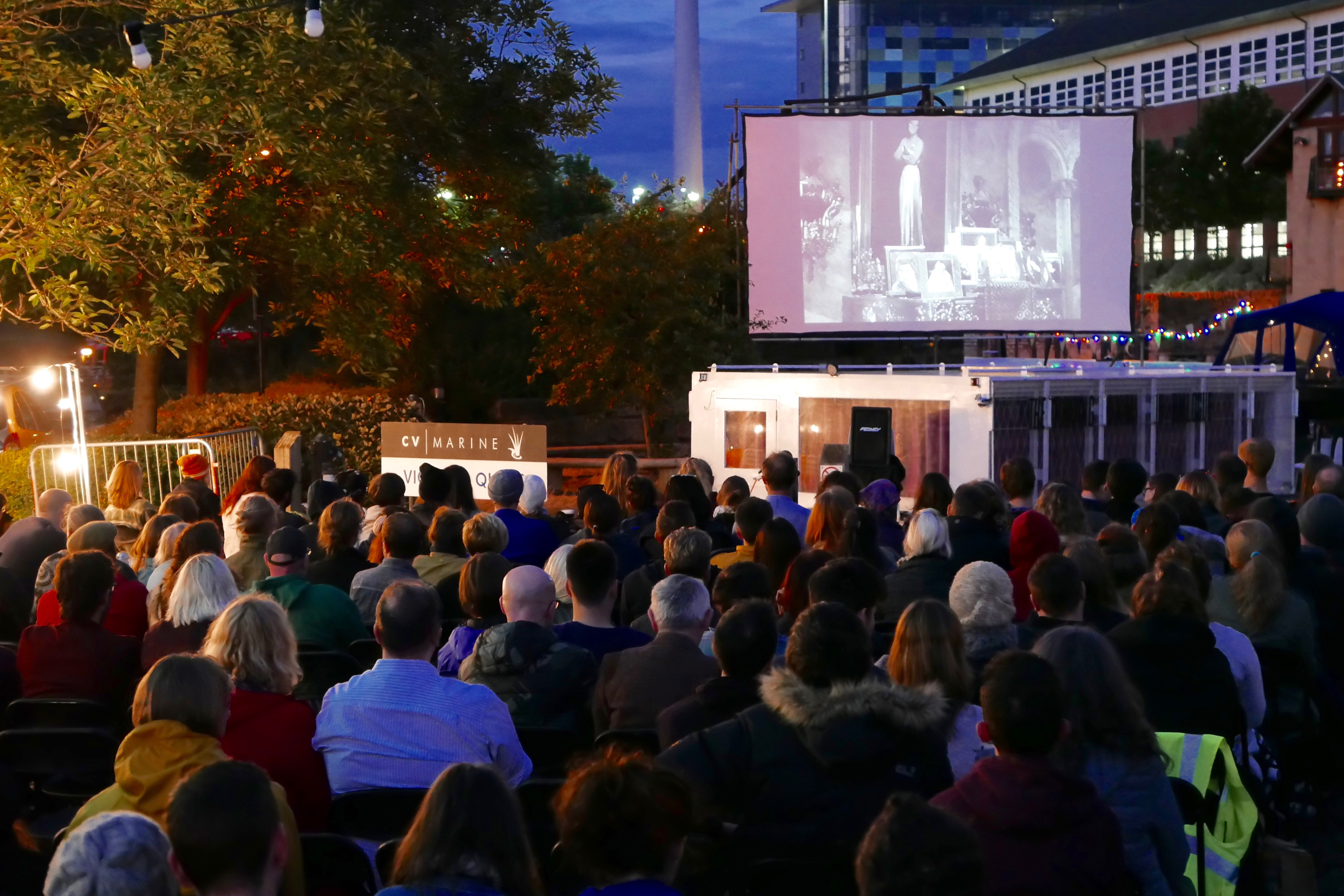 A large group of people at night all sitting on chairs facing towards a projection screen, with hanging lights all surrounding them.