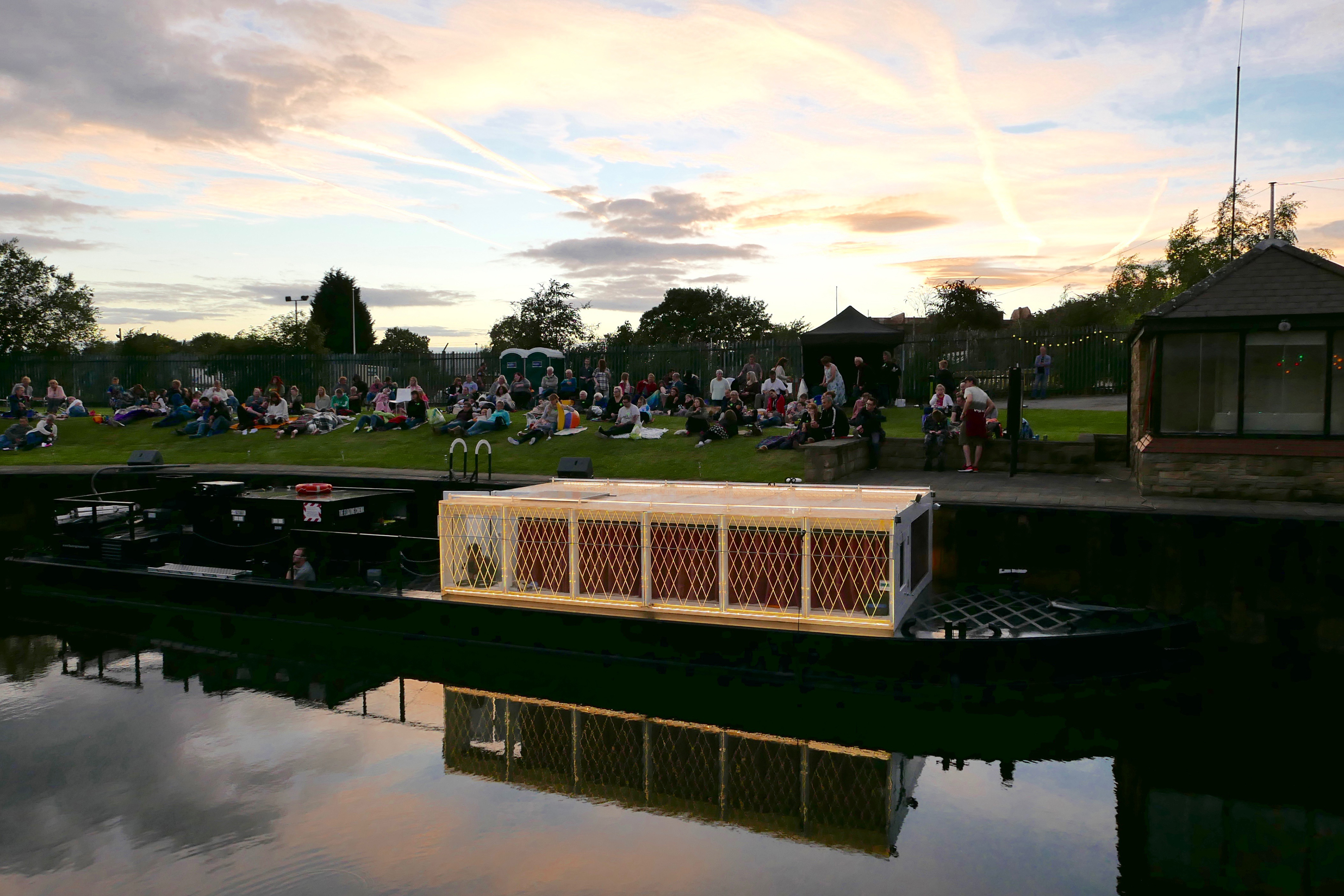 A large white barge canal boat at dusk with lights on, next to a bank of grass full of people sitting down.