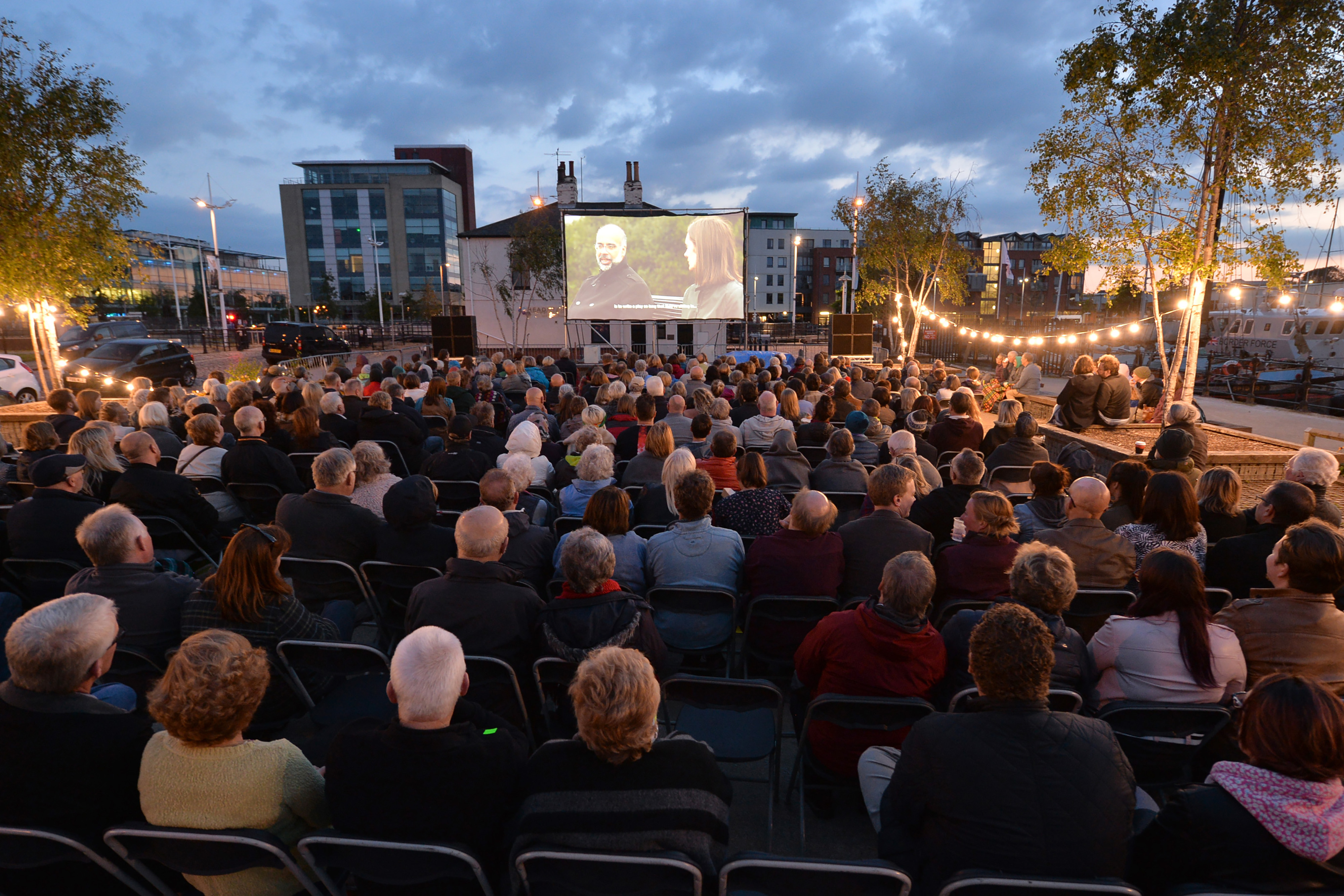 A large group of people at dusk all sitting on the ground facing towards a projection screen, with hanging lights all surrounding them.