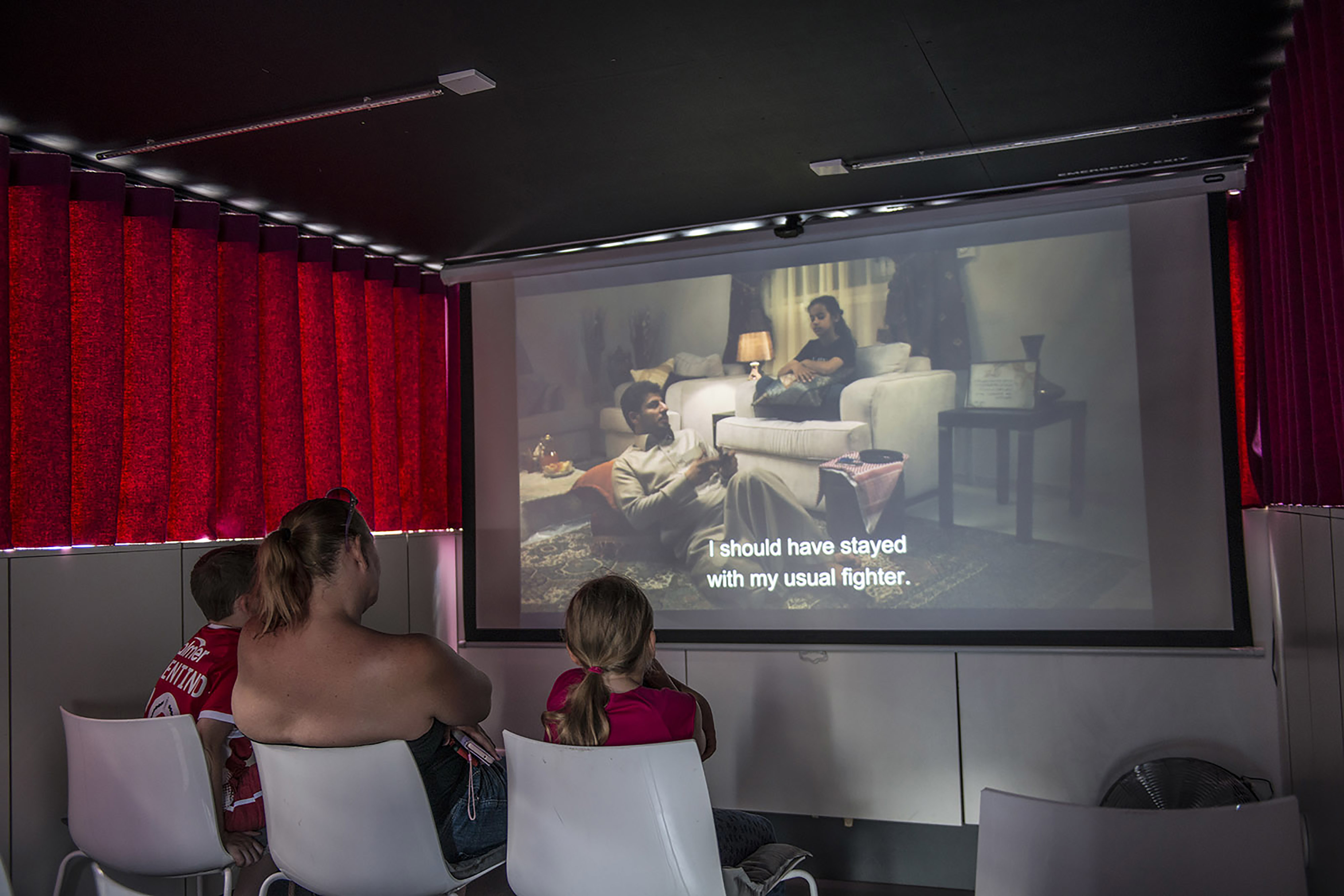 A woman and two young children in a small room with purple curtains all watching a small projection screen showing a film.