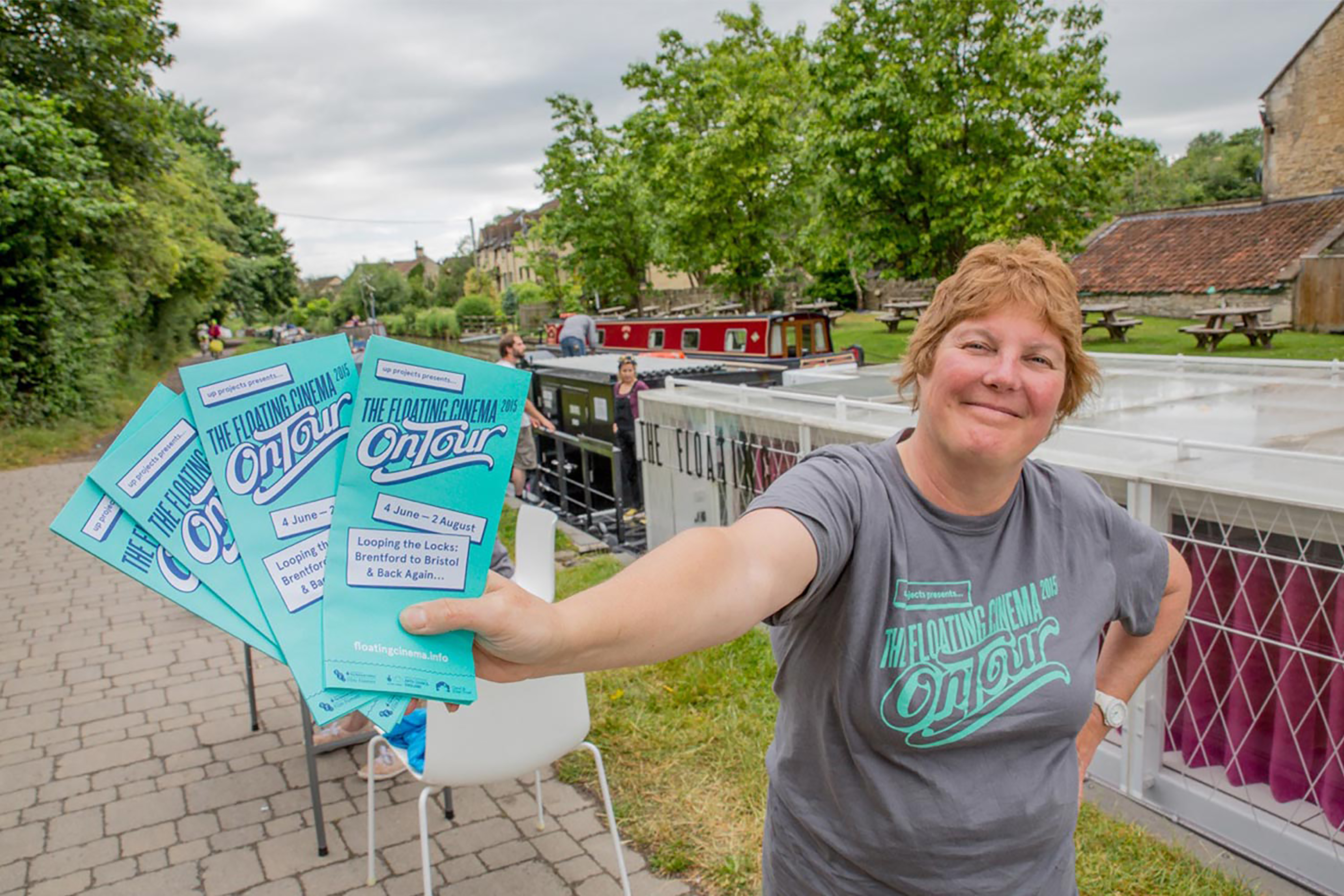 A woman holding her arm out holding blue flyers, standing in front of a large white canal boat on a river.