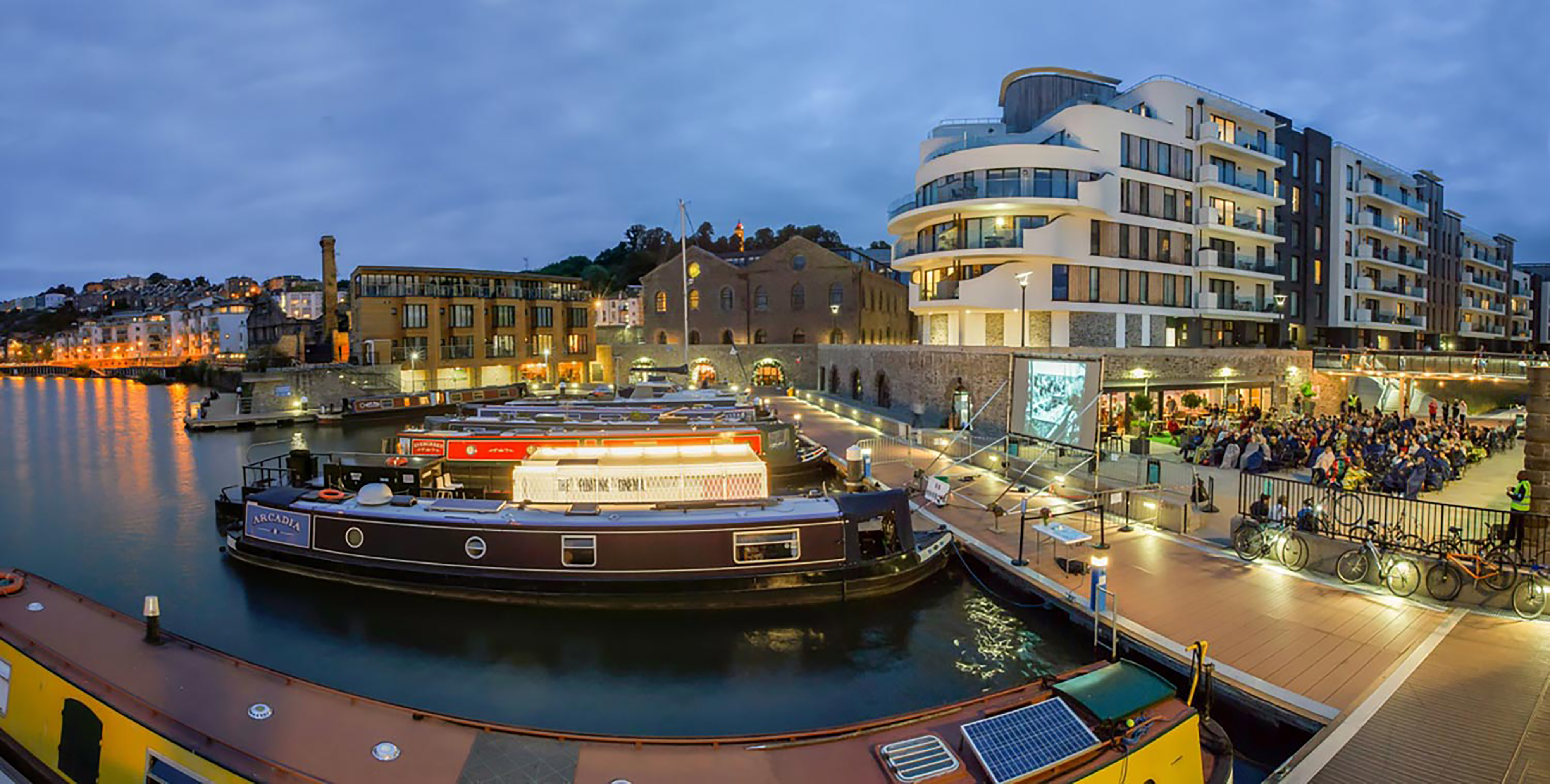 A panoramic photograph of a boating dock at night, with several boats, in front of a large apartment block and group of people in the background.