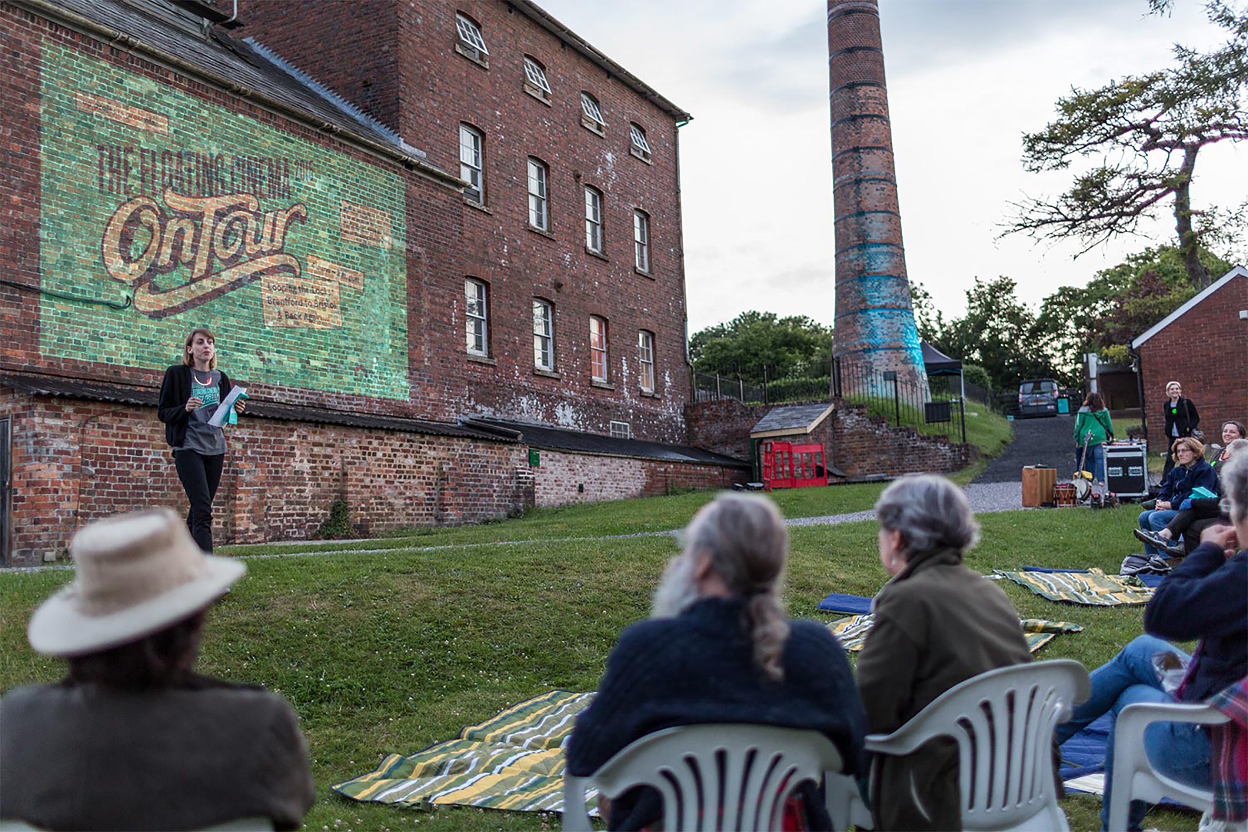 A group of people sitting on plastic garden chairs all watching a person speaking in front of a large old red-brick building with an image projected onto the side of the building.