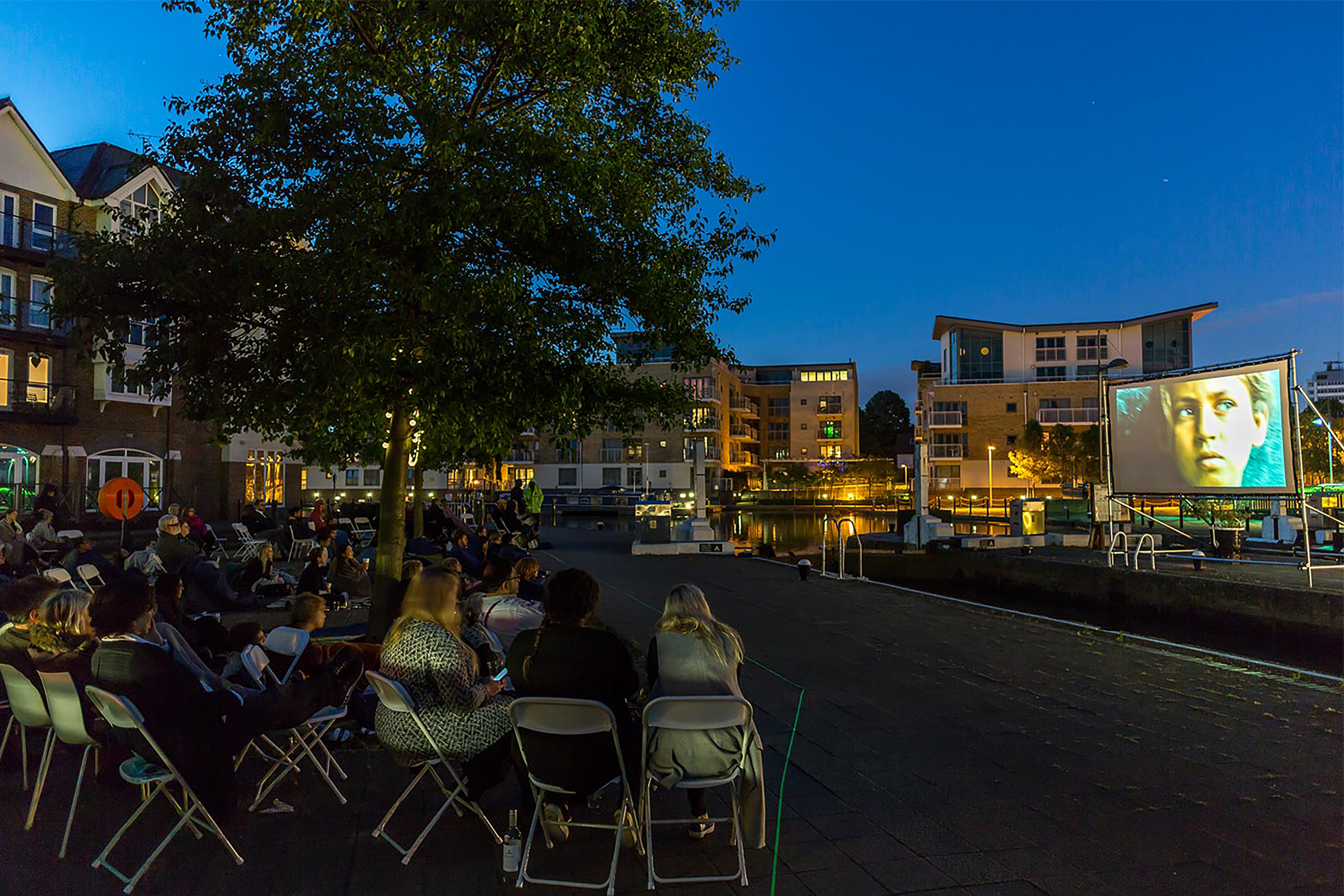 A large group of people at dusk all sitting on the ground facing towards a projection screen.