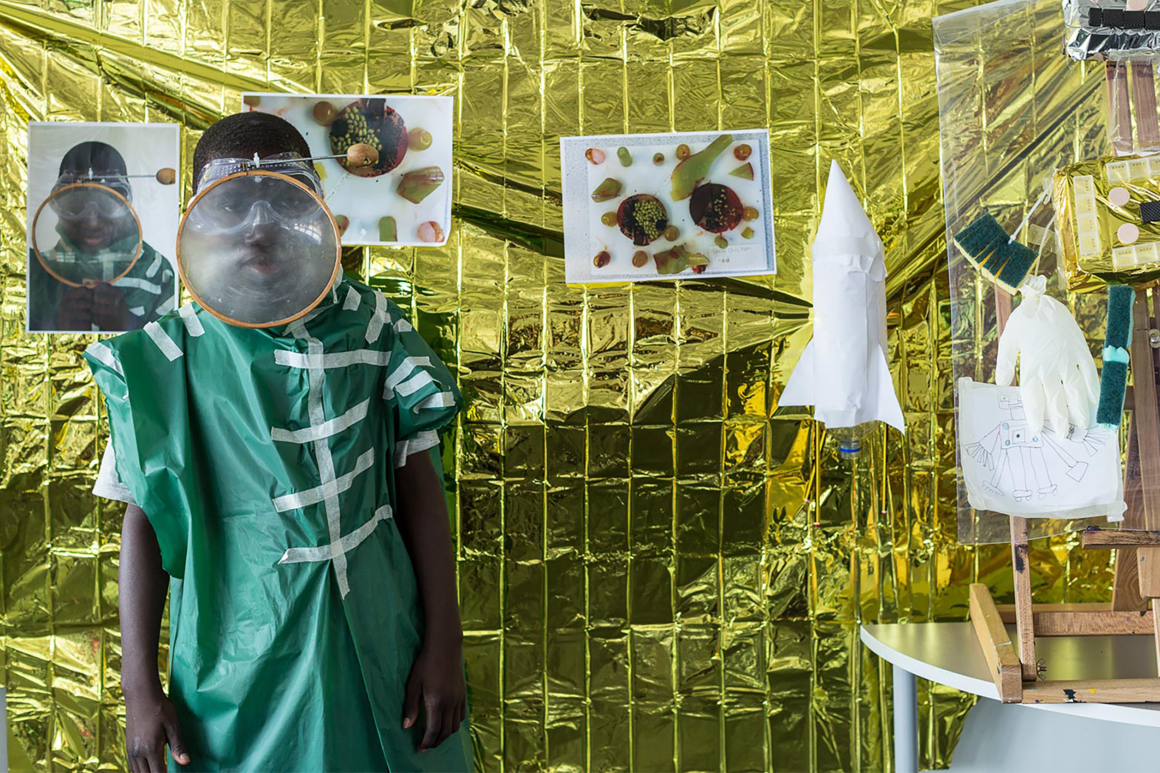 A black boy standing in front of a gold foil backdrop with pictures on it, wearing a green handcrafted garment and magnifying glass on his face.