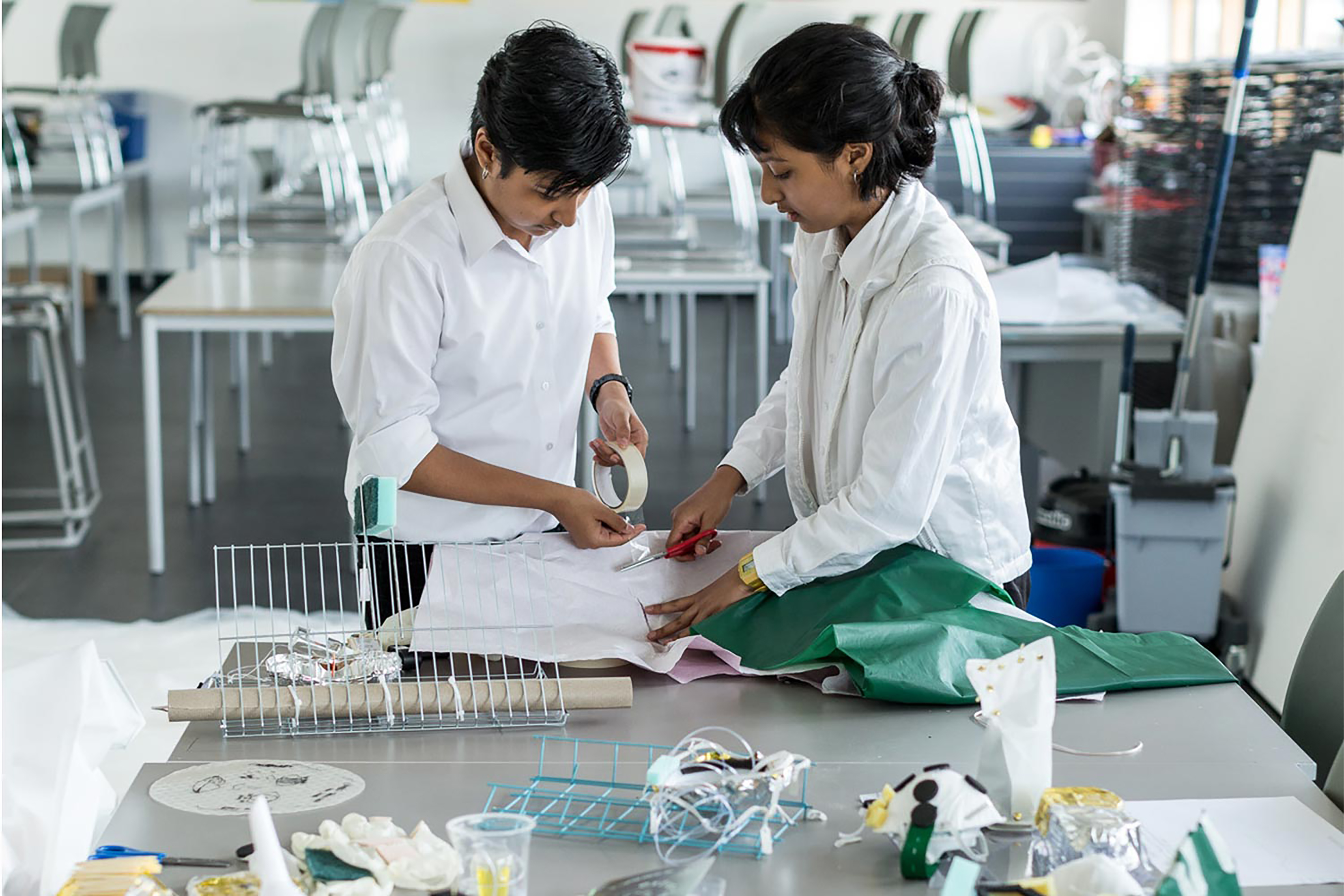 Two South Asian school children wearing white shirts handcrafting items in an untidy workspace full of tables and stackable chairs.