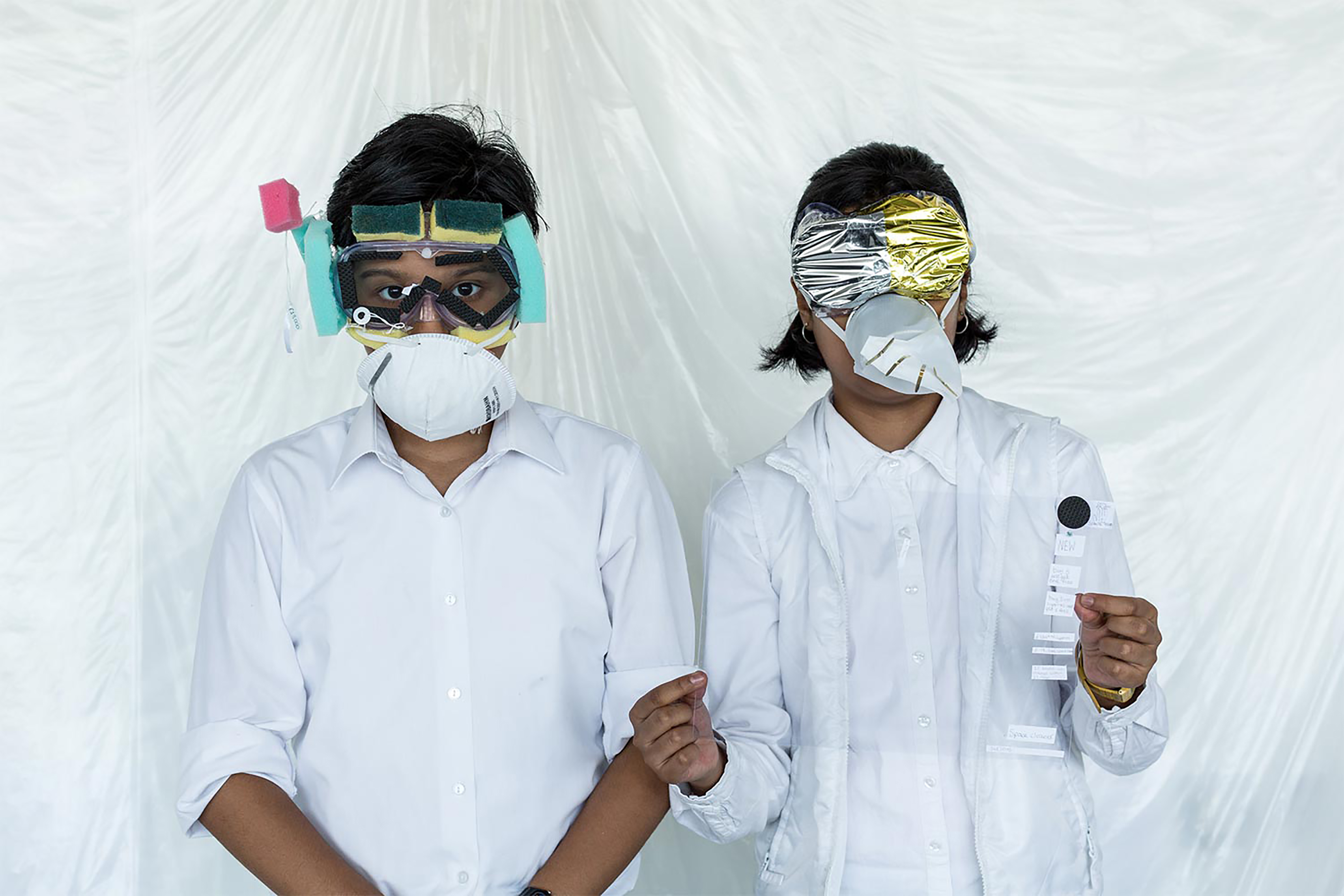 Two South Asian school girls wearing white shirts and handcrafted face masks standing in front of a clear white sheet background.