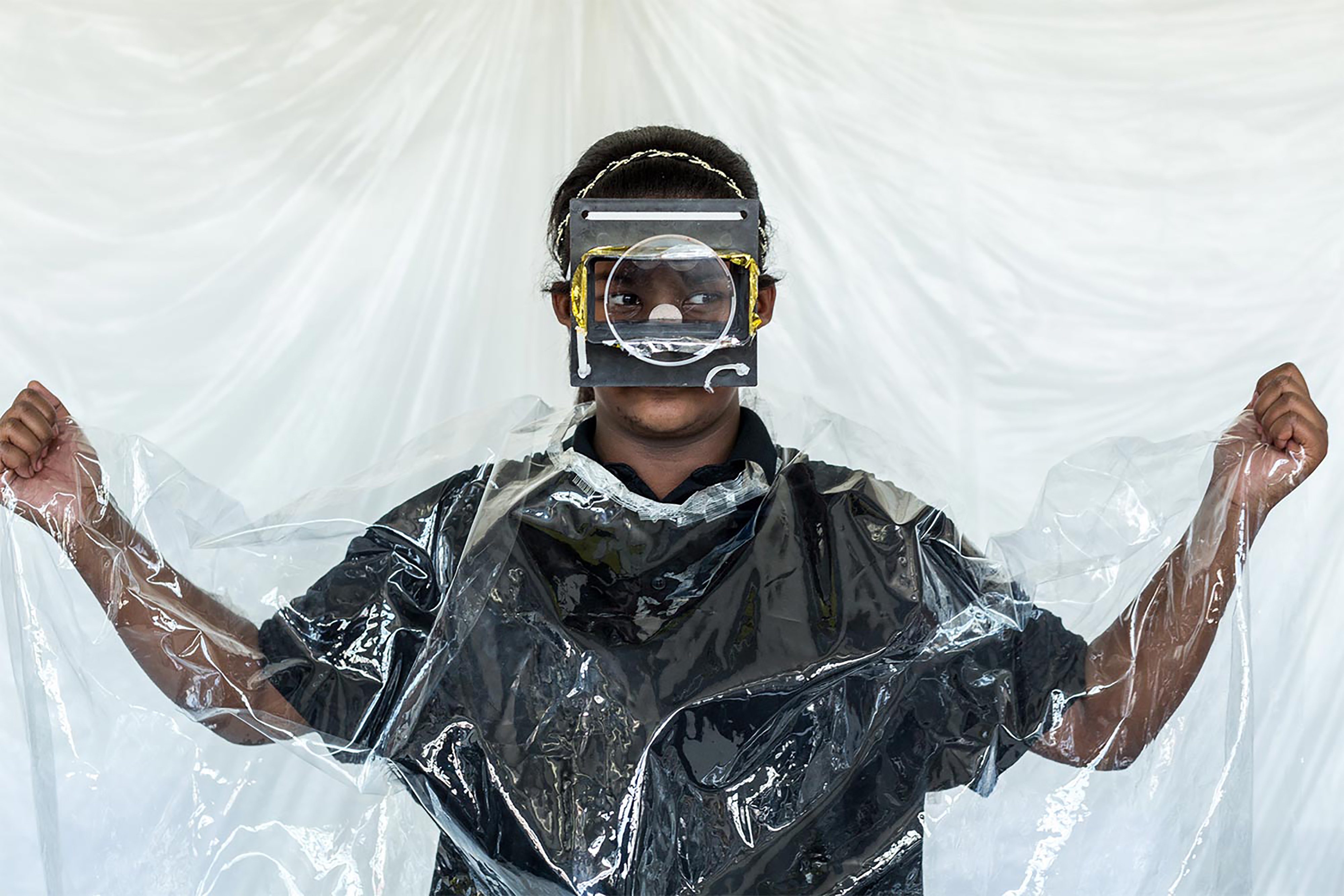 A South Asian school child wearing a black shirt, a transparent disposable rain poncho, and a handcrafted face mask in front of a white sheet background.