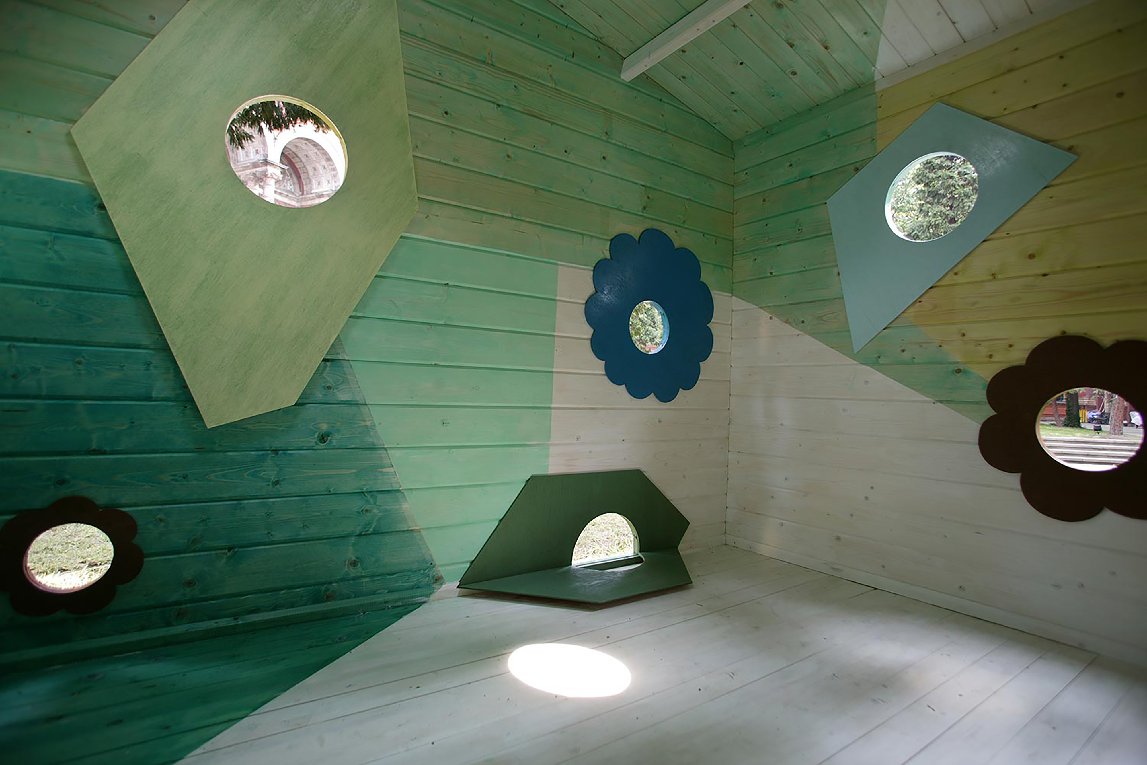 Inside of a garden shed with wooden interior and walls painted in a geometric form in various shades of green, with 3d shape panels overlaying small gaps of circular windows letting the light in.