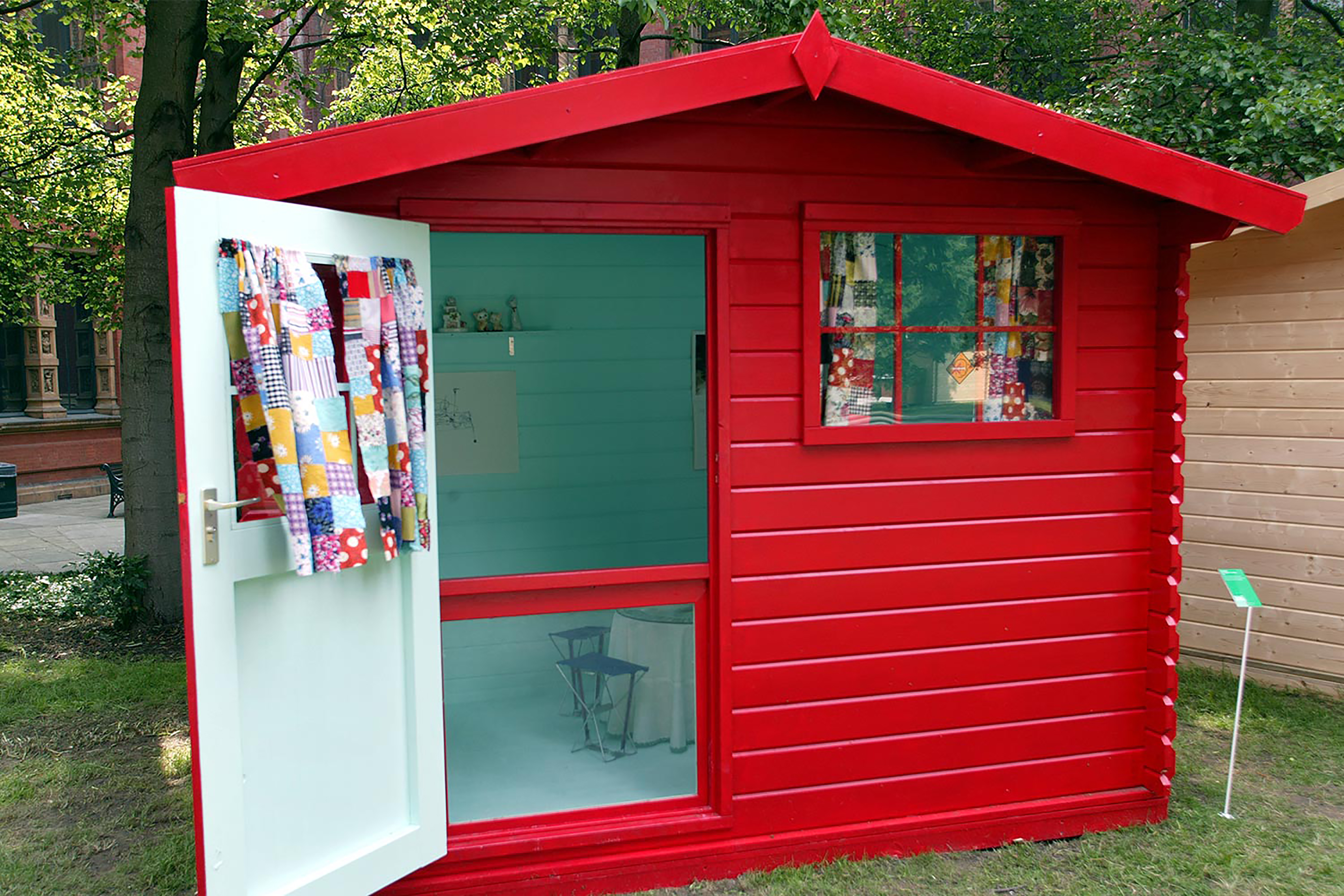 A bright red wooden garden shed on a patch of grass, with a blue painted wall inside and a colourful patchwork fabric hanging on the door.