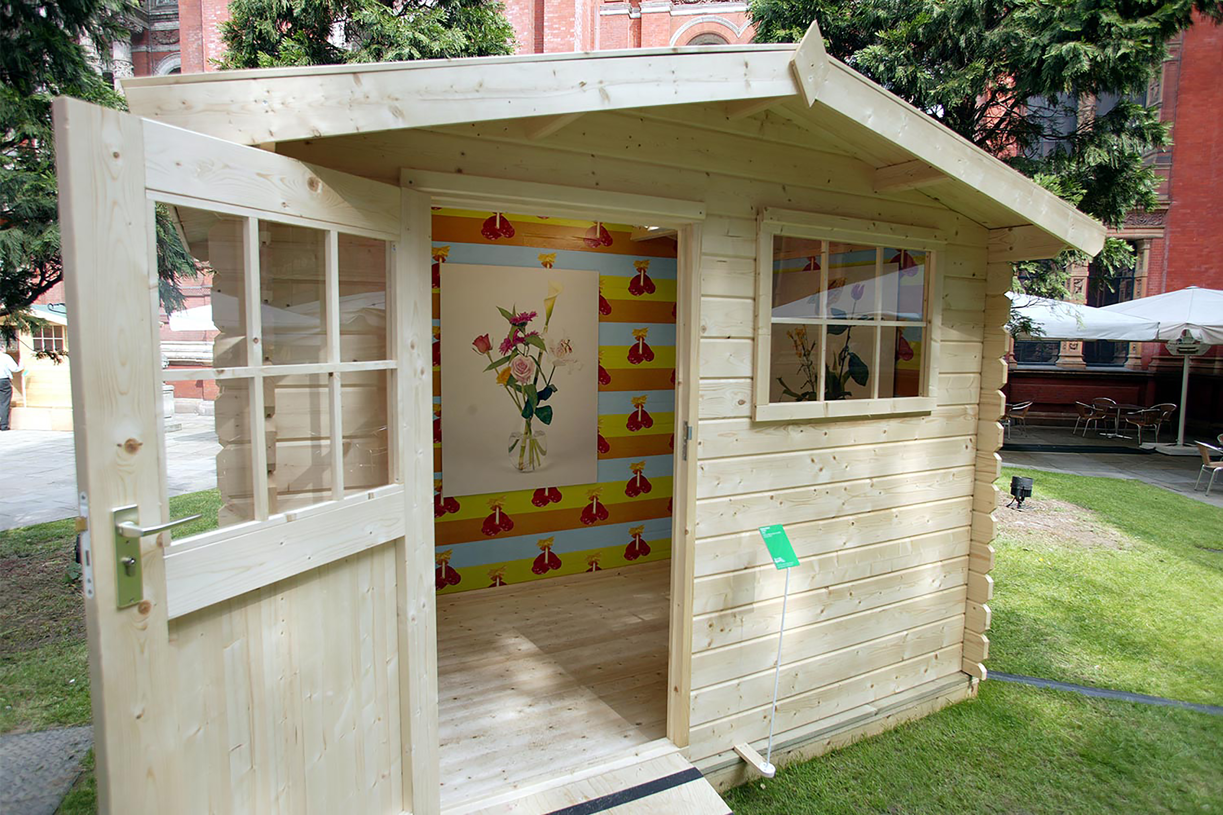 A brown wooden garden shed on a patch of grass, with colourful wallpaper print inside and a picture of a botanical flower hanging up.