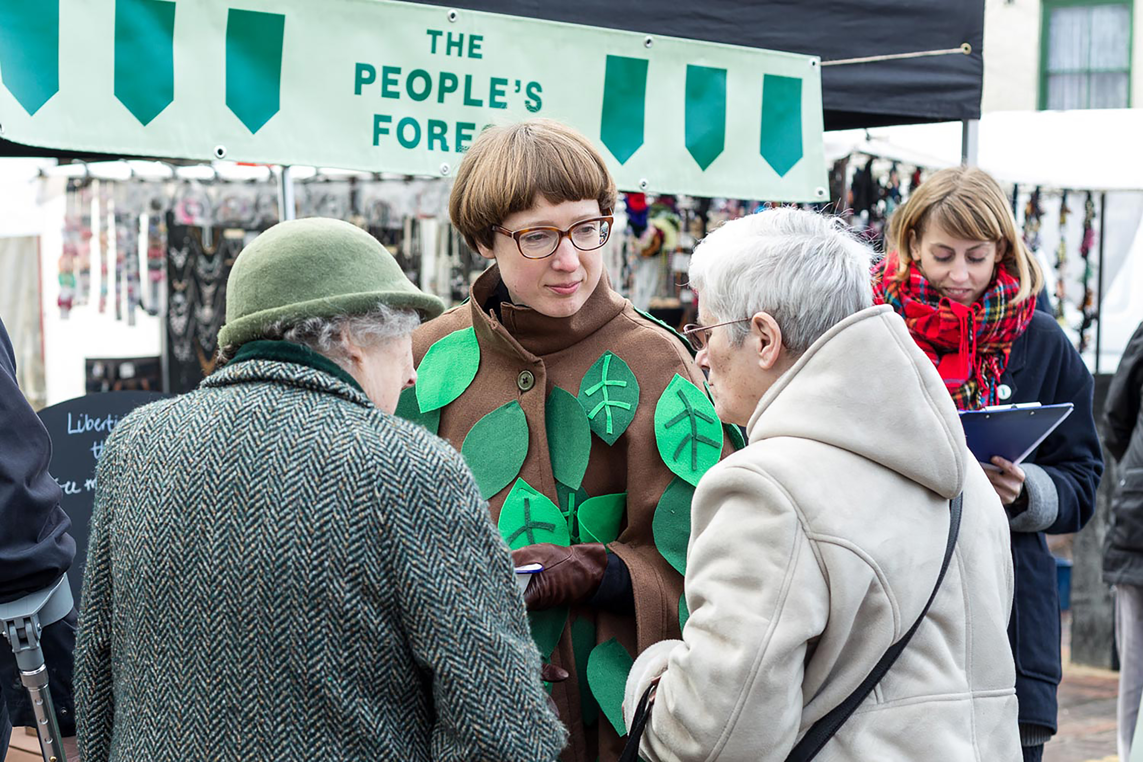 A white woman wearing glasses and a leaf covered coat holding a clipboard while speaking with two elderly white women in front of a green banner with the words “The People’s Forest”.