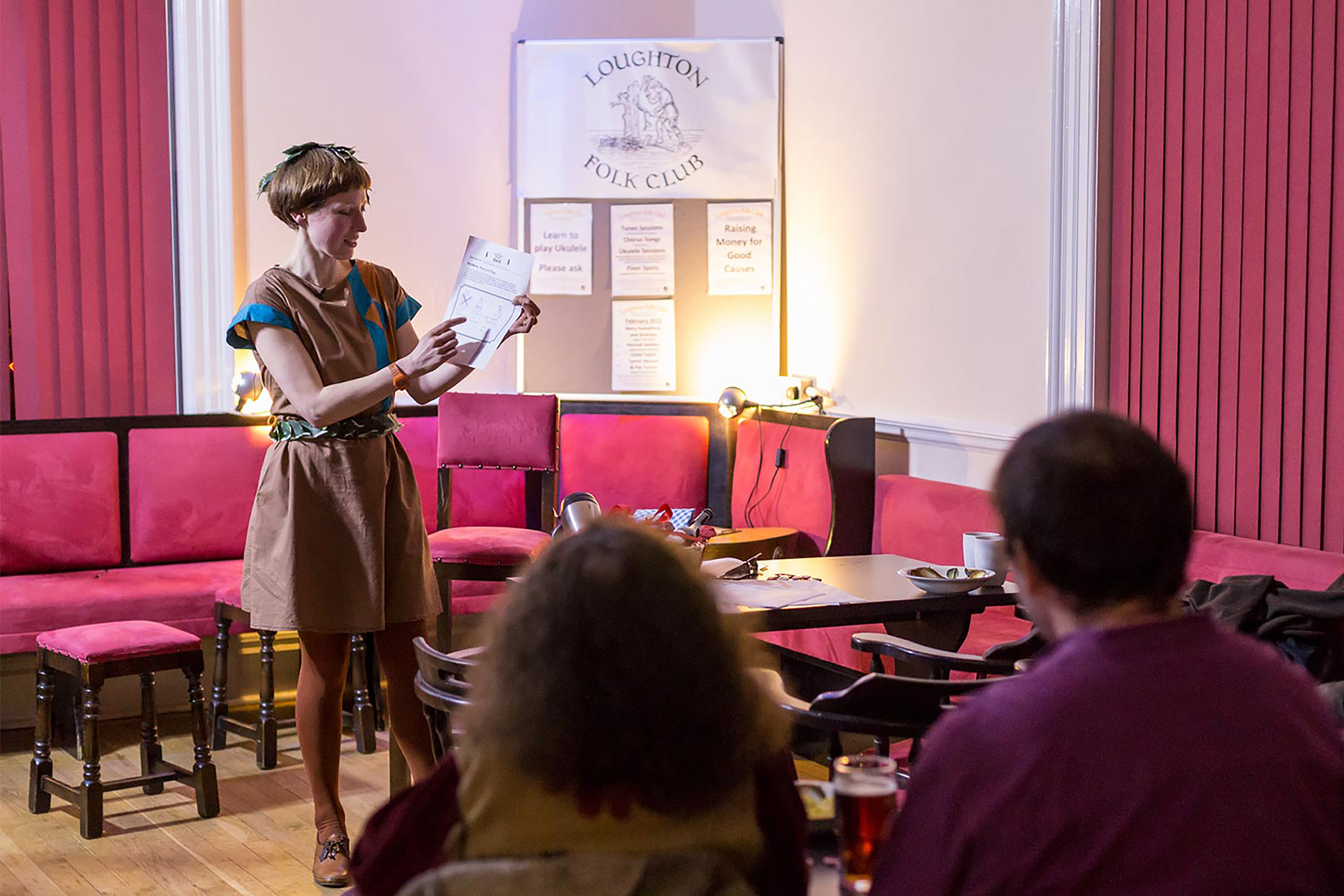A white woman wearing a green and brown tunic holding a sheet of paper, in front of a crowd gathered in a social club with dark pink chairs and curtains.