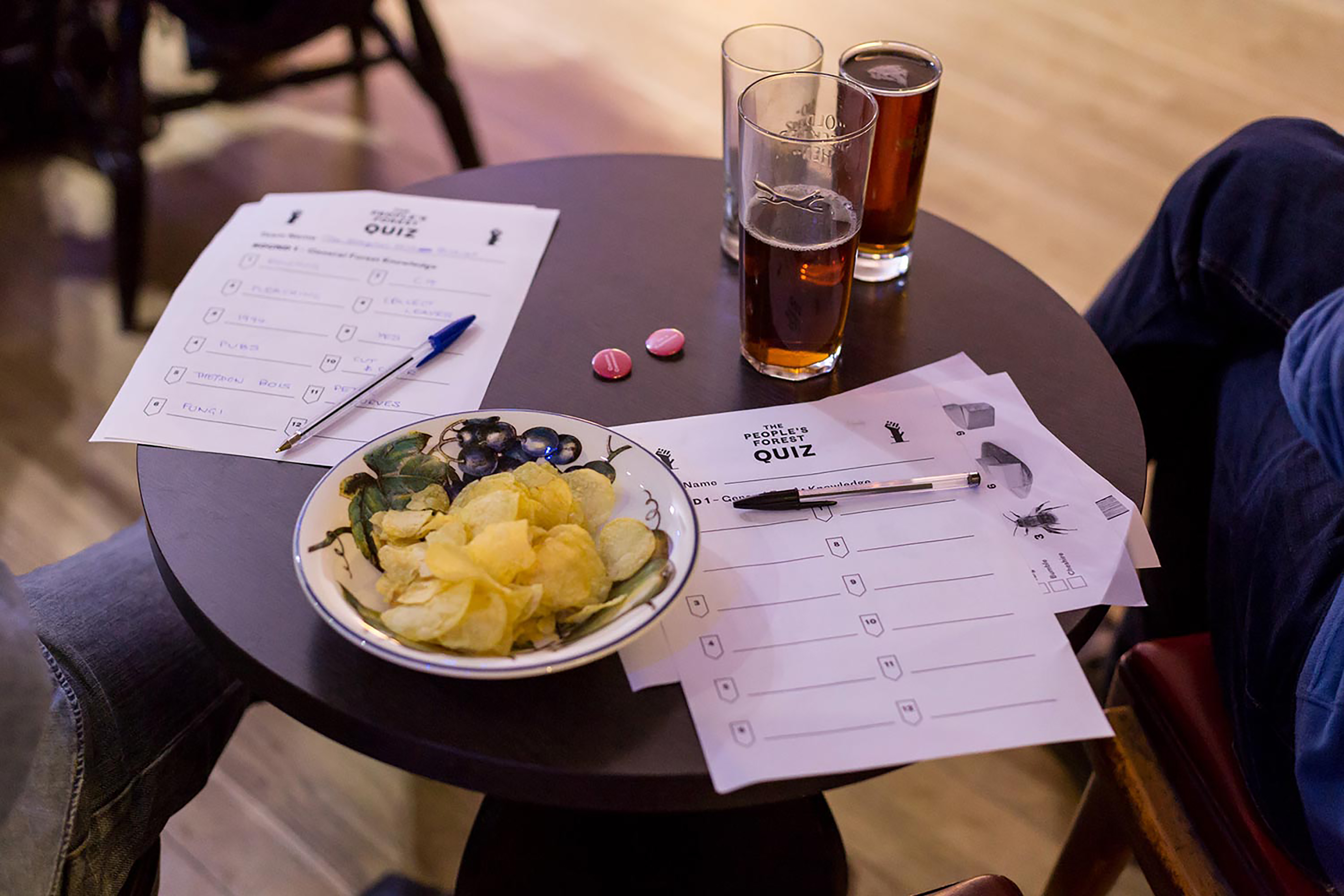 A bowl of crisps, three pint glasses and several A4 quiz sheets on top of a brown circular wooden table.