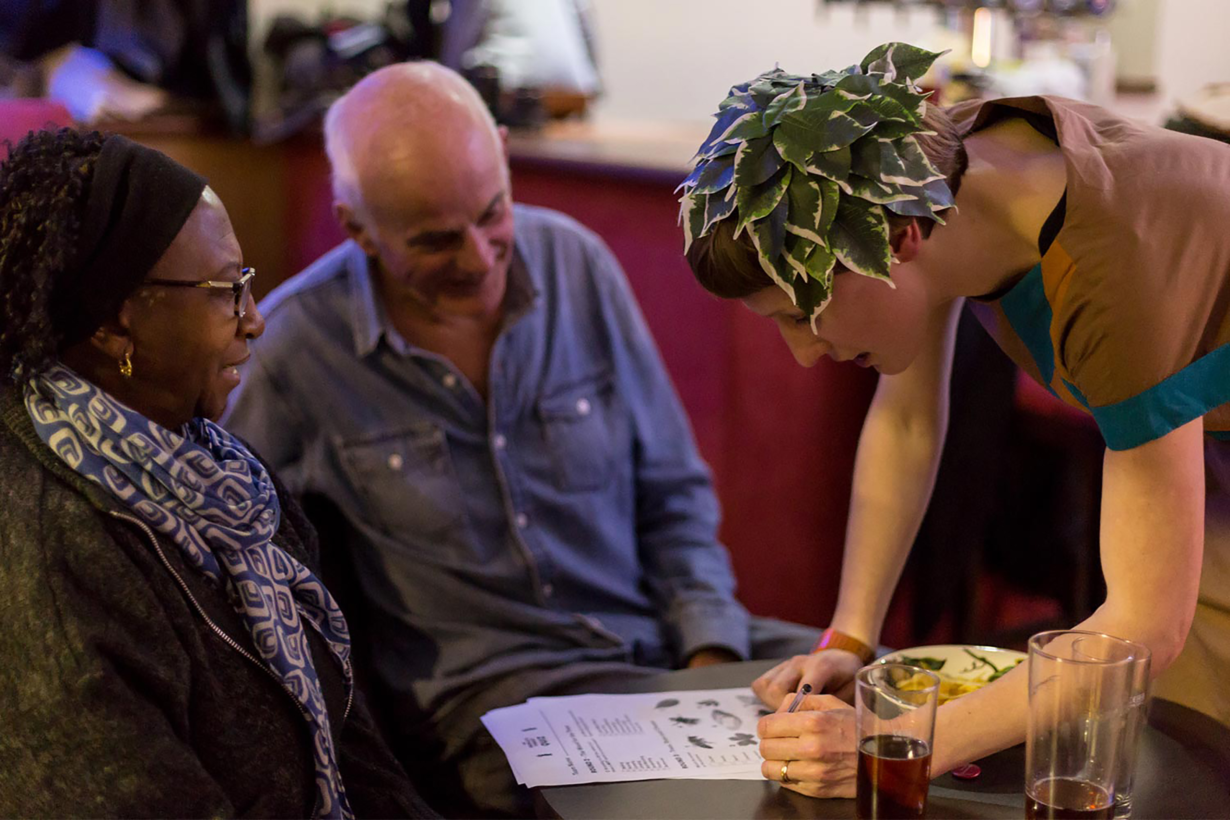 A white woman wearing a tunic and a leaf headdress marking an A4 quiz sheet on a table while an elderly man and woman sit and observe in a social club.
