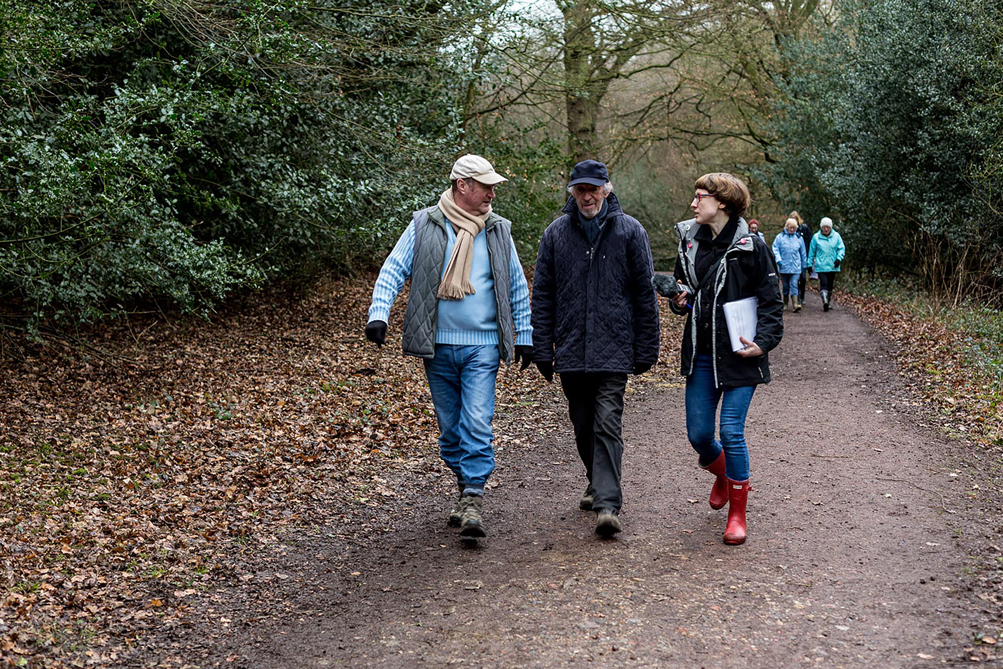 Three people walking down a path in a woodland area, all wearing raincoats and wellies.