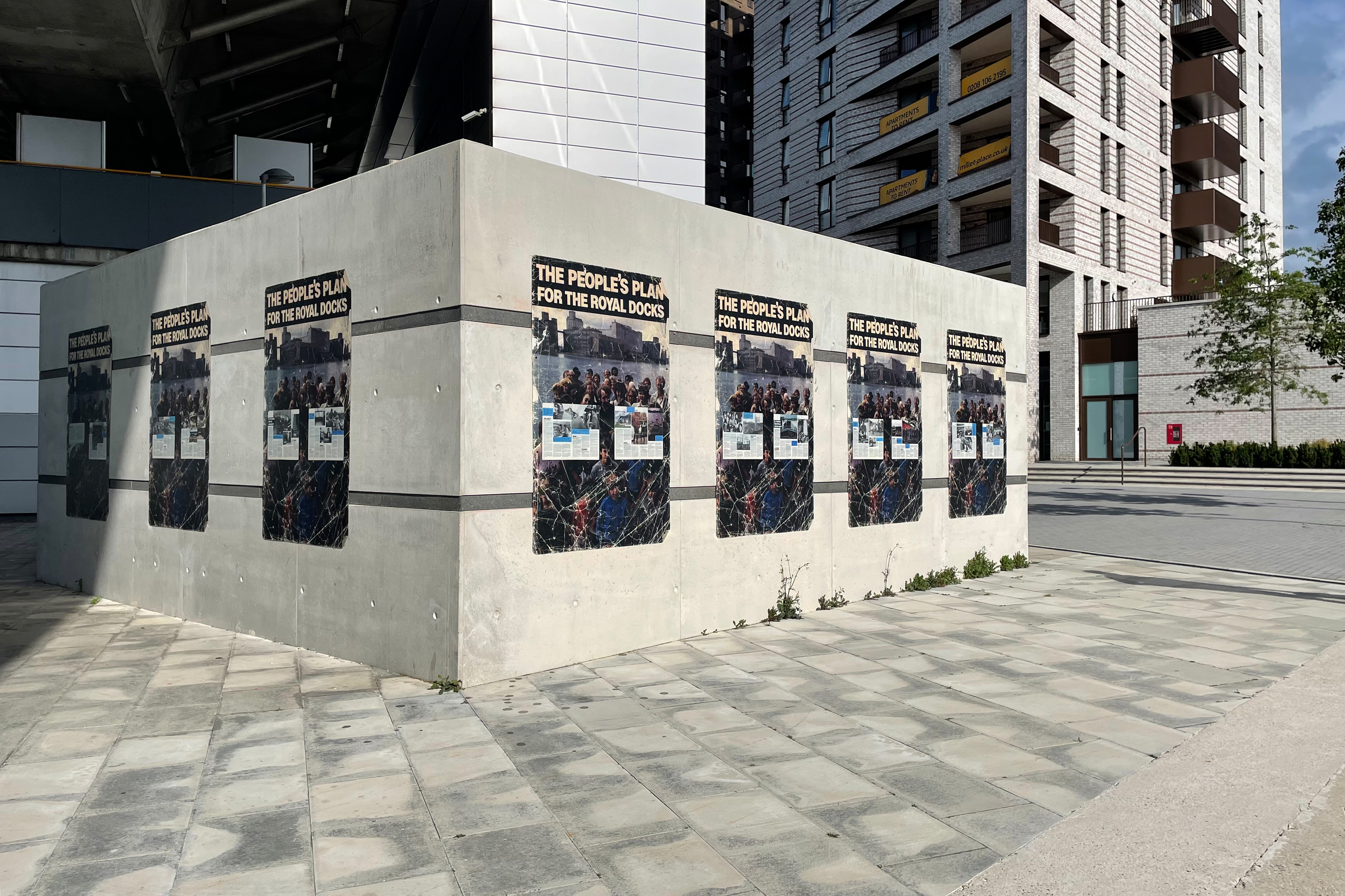 A large square concrete block covered in artwork prints with images of historical residents, the words "The People's Plan for the Royal Docks", all spaced out in front of white apartment buildings.
