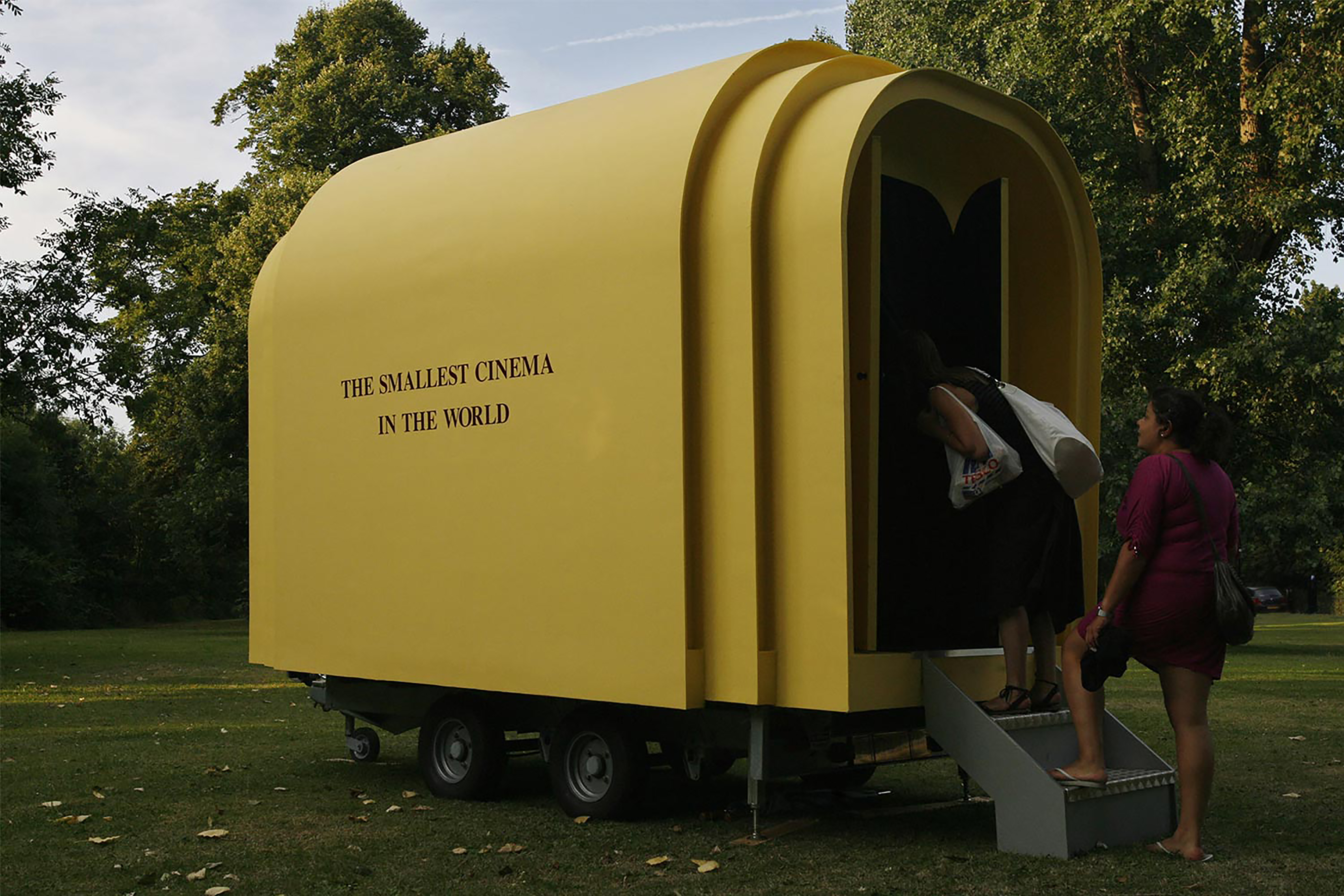 A small yellow outdoor cinema pod with the words "The Smallest Cinema in the world" on the side, with people climbing inside.