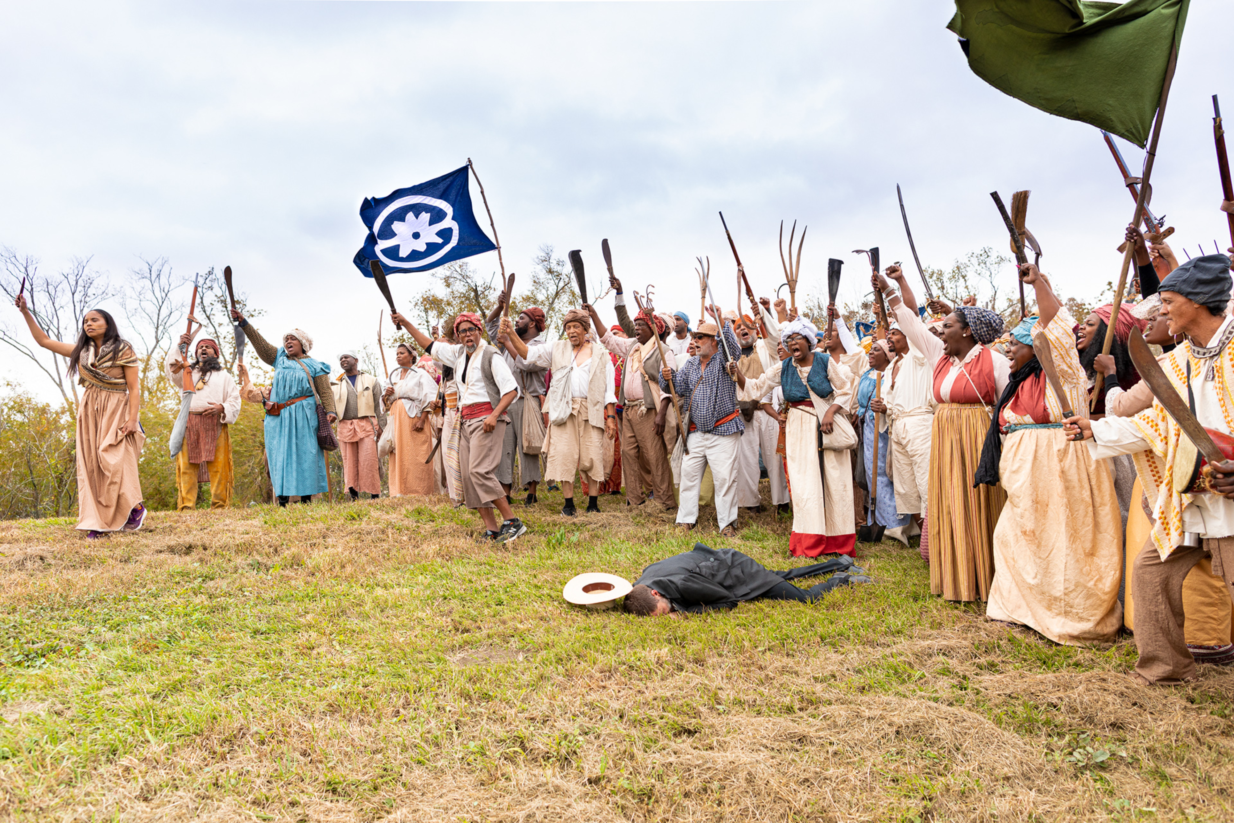 A large group of people dressed in period clothing. They are standing in a line, chanting and holding various objects in the air.