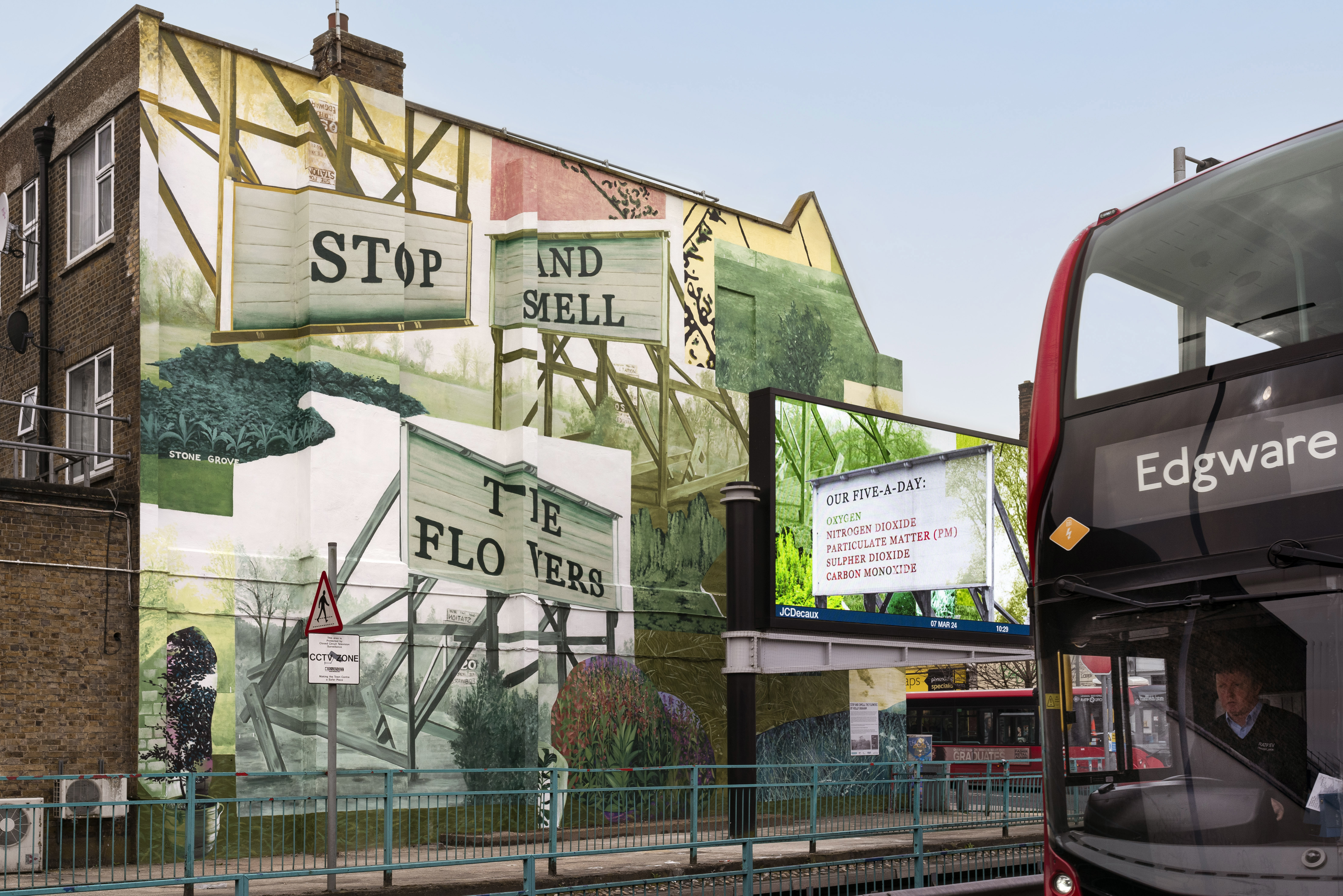 A painted mural on the side of a large building’s wall. The artwork has different layered pictures of trees, plants and nature. The words include "Stop and smell the flowers" which are presented next to a digital screen with a London red bus driving past.