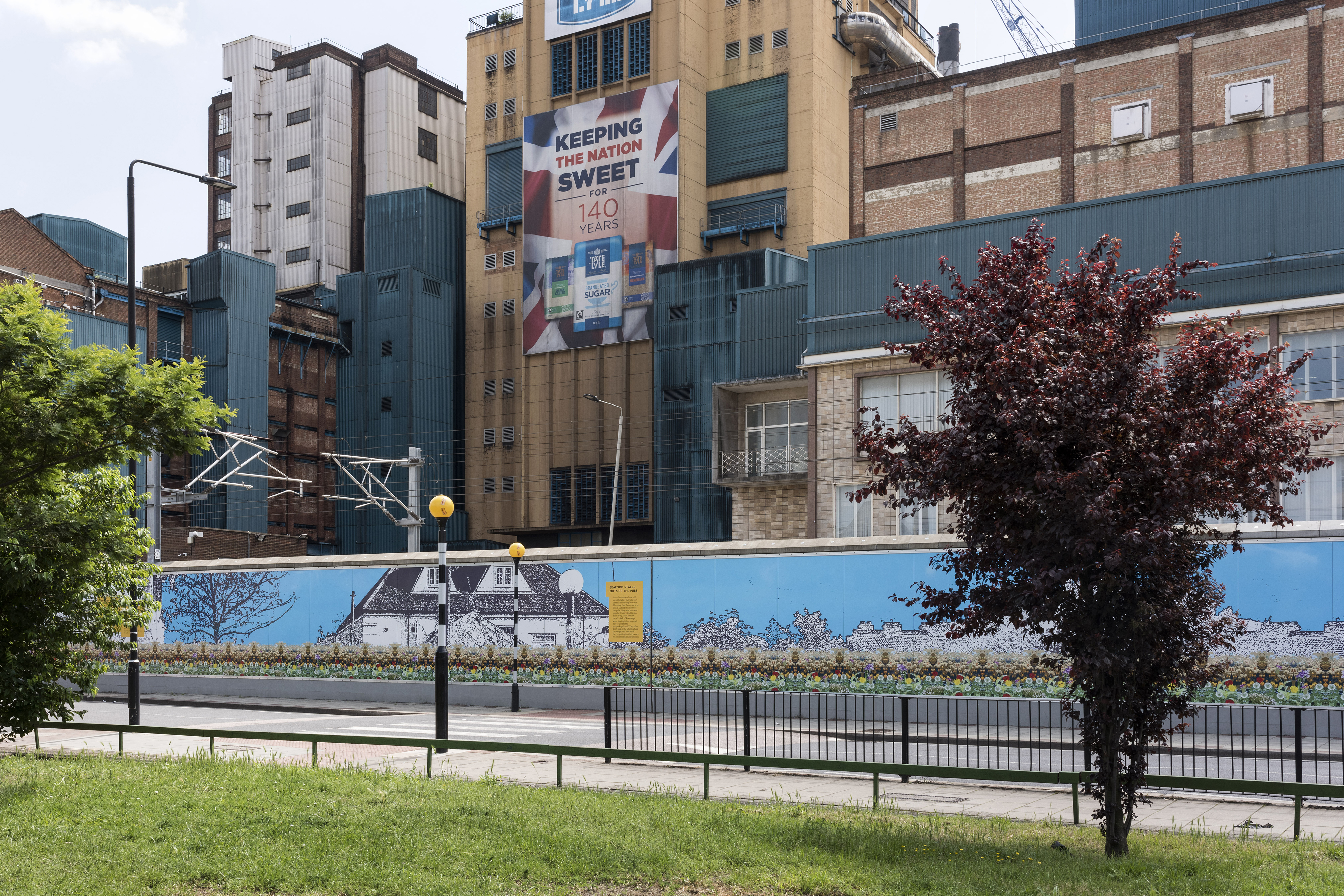 A photograph of an empty road accompanied by a zebra crossing and a trackside wall covered in artistic murals in front of a railway track and sugar factory.