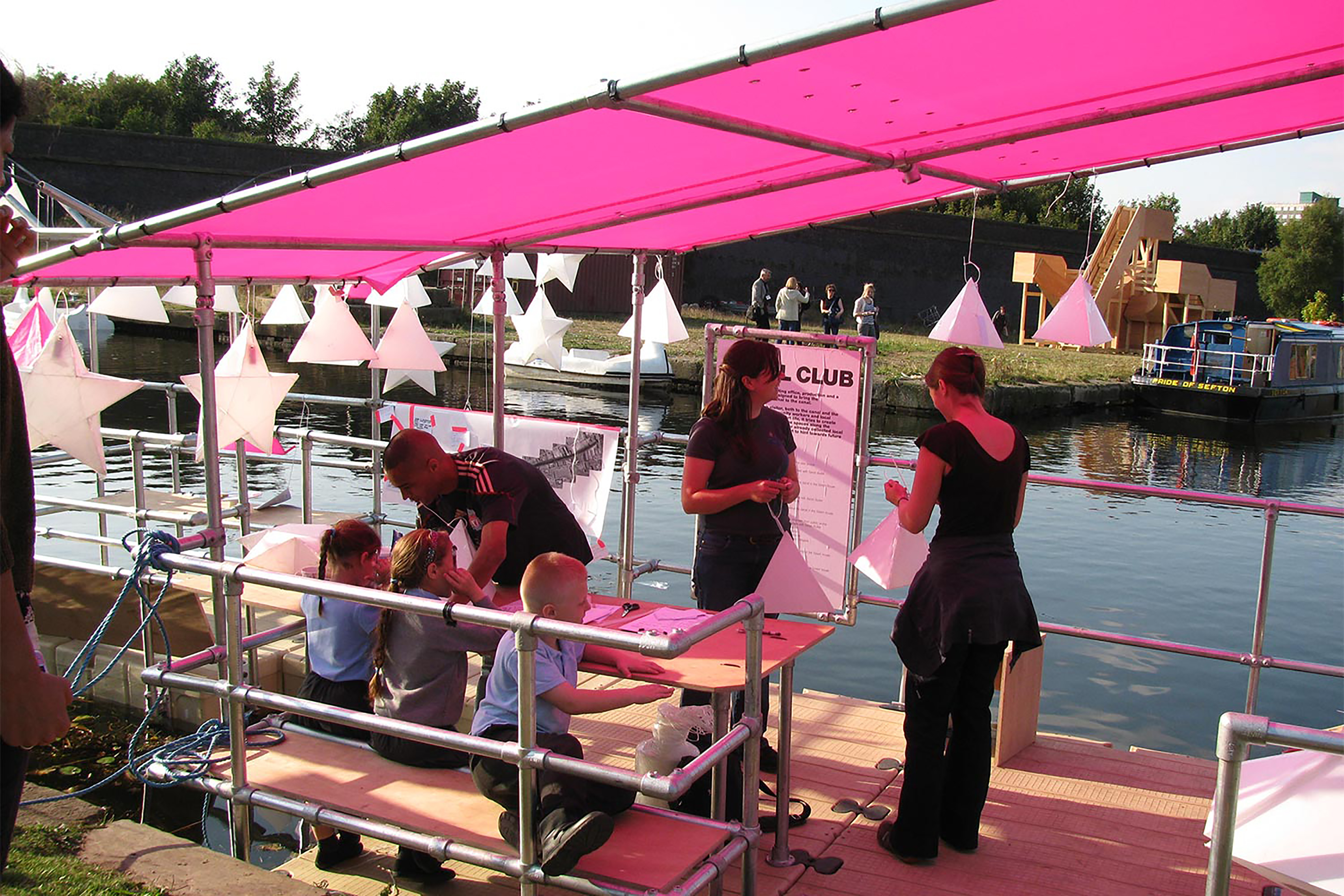 A photograph of children and adults creating paper pyramids on a dock, under a large pink canopy.