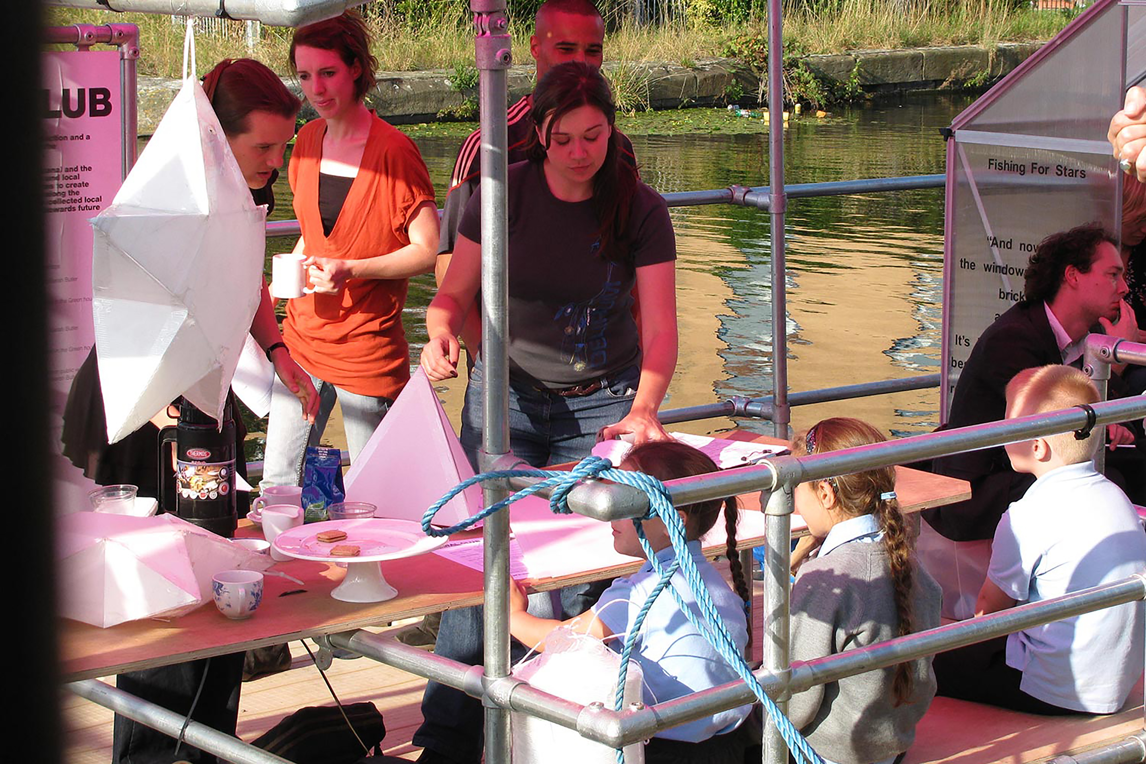 A photograph of children and adults creating paper pyramids on a dock, surrounded by pink posters and metal scaffolding.