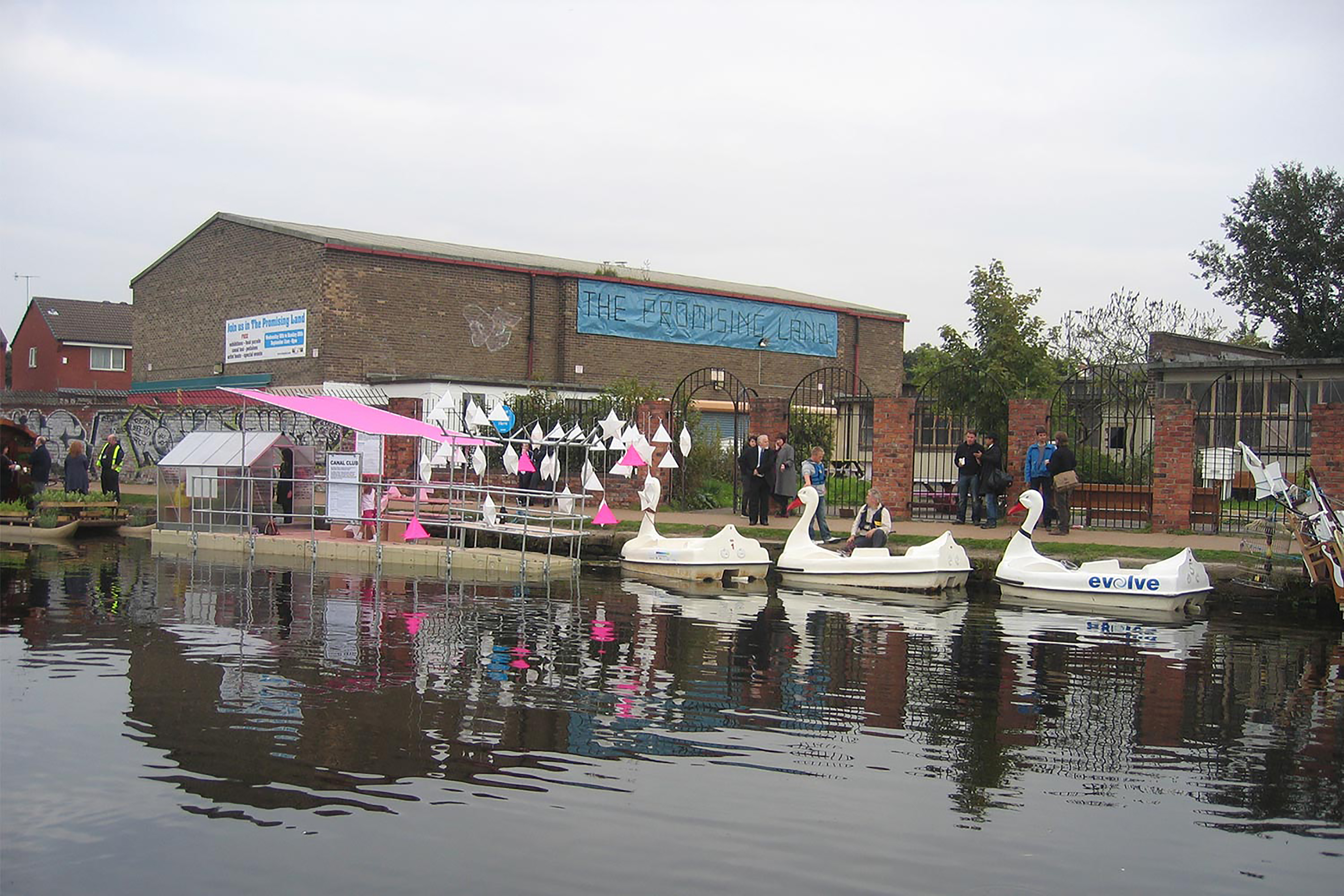 A photograph of a canal dock and three swan pedalos, next to a bank fill of people.