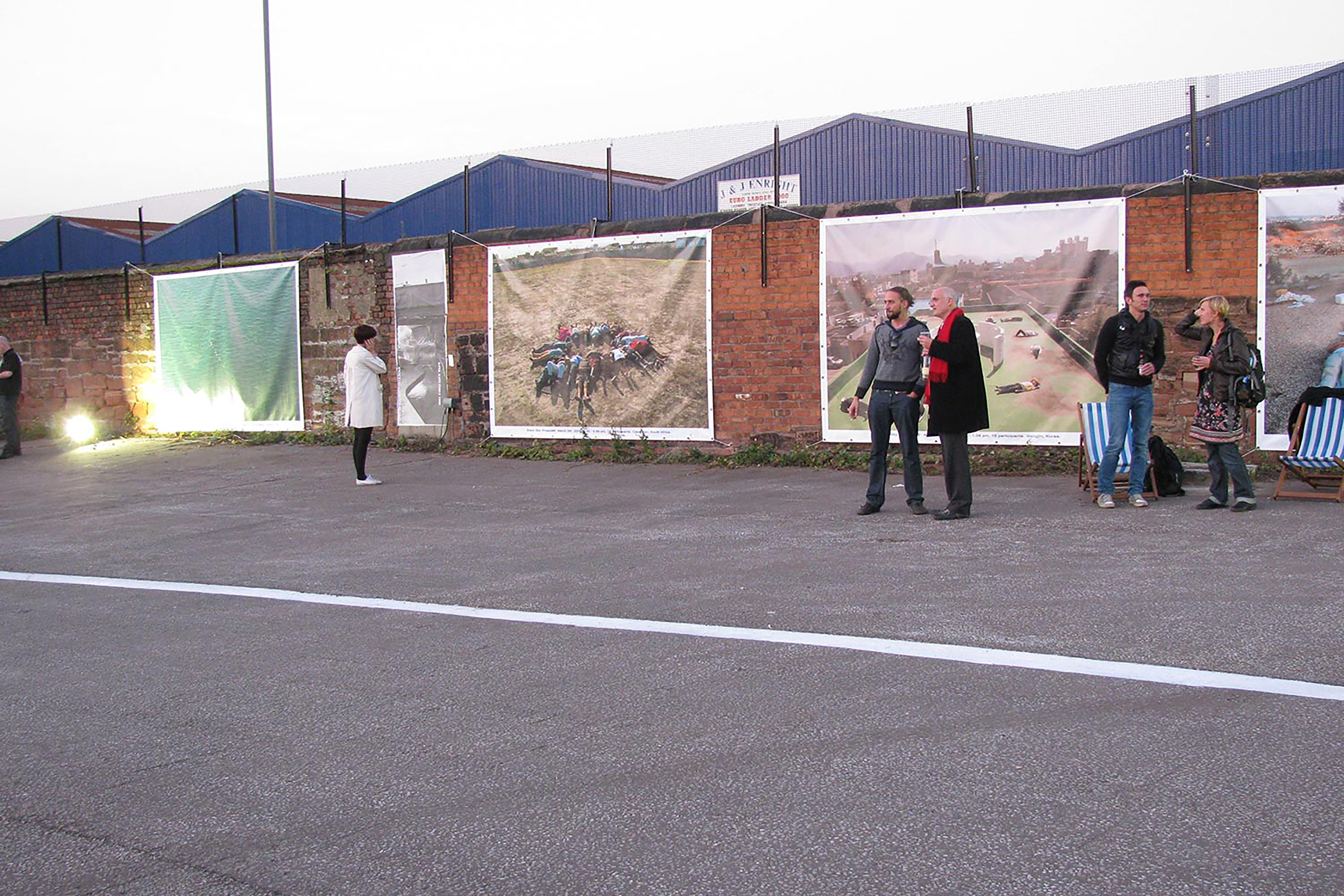 A photograph of a few adults gathered around a brick car park wall covered in large photos.