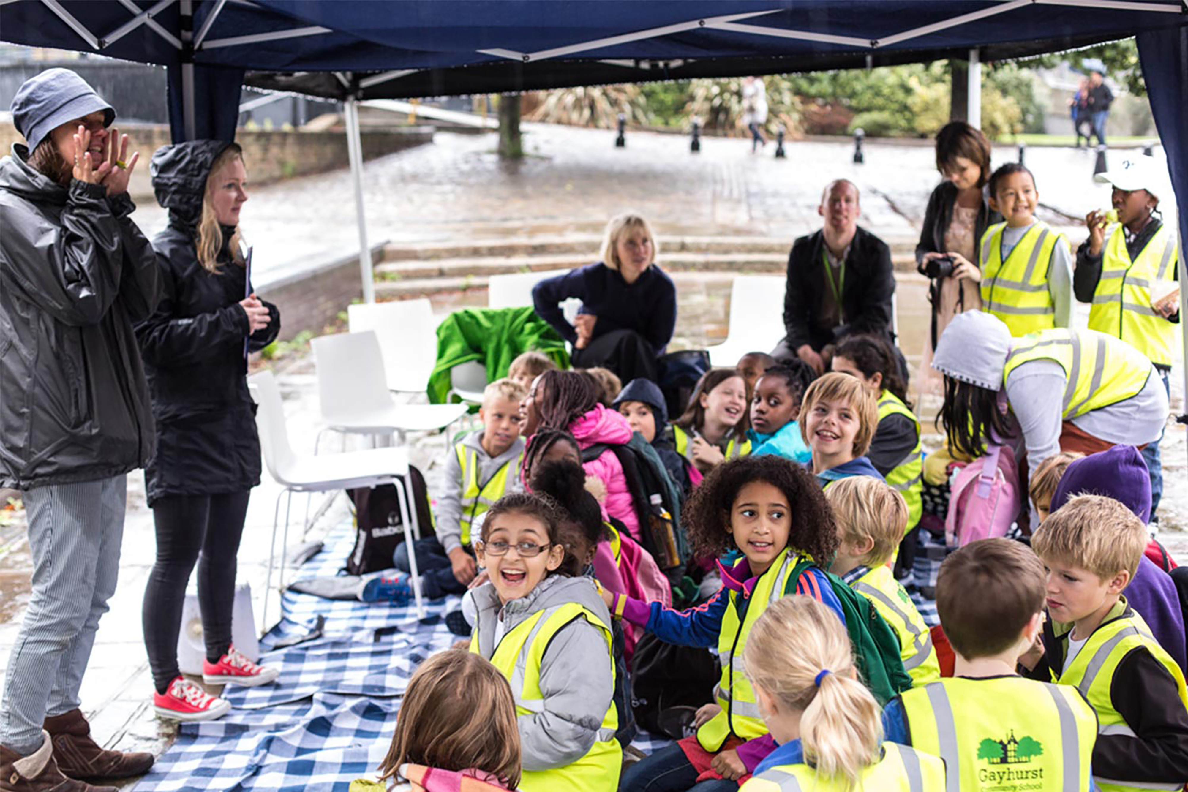 Two women wearing black raincoats talking a crowd of children wearing green Hi-Vis jackets sat under a gazebo.