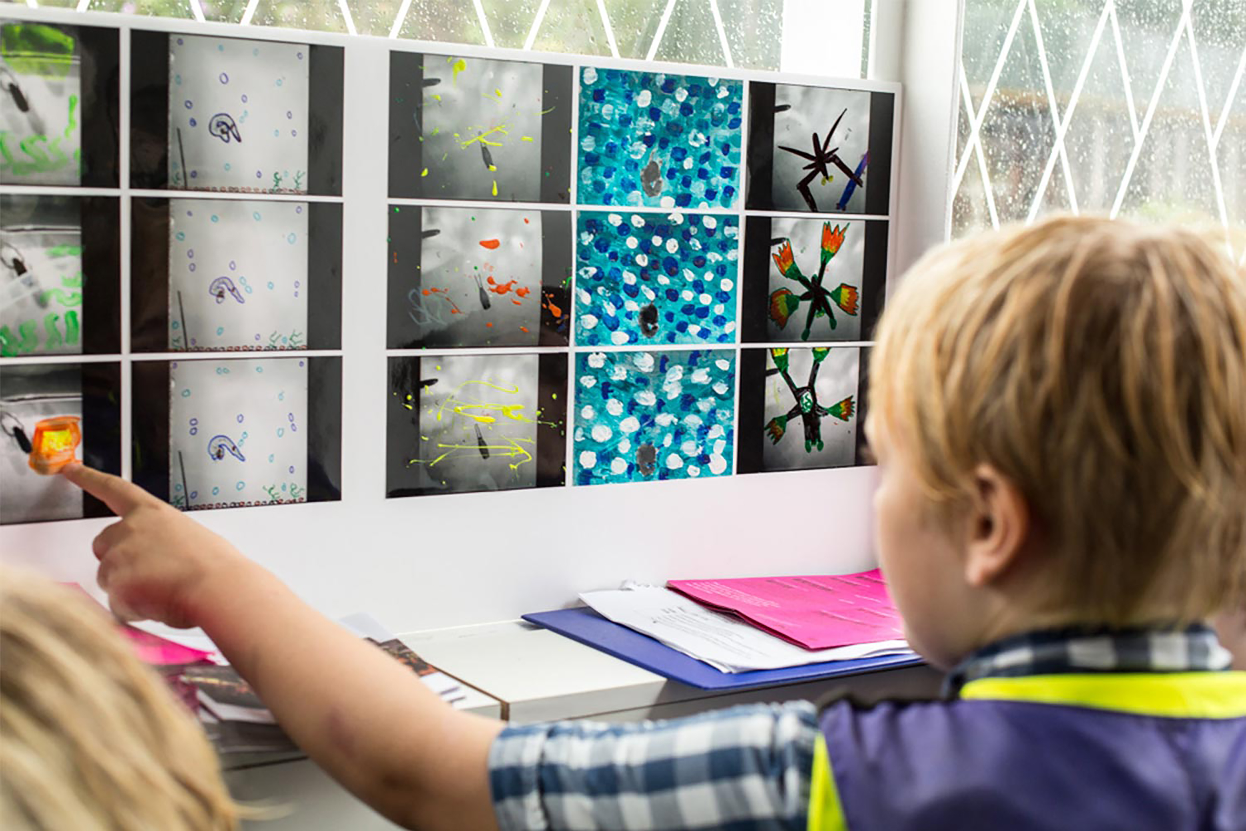 A young boy wearing a green Hi-Vis jacket and check shirt pointing to a collage of paintings stuck to a whiteboard.
