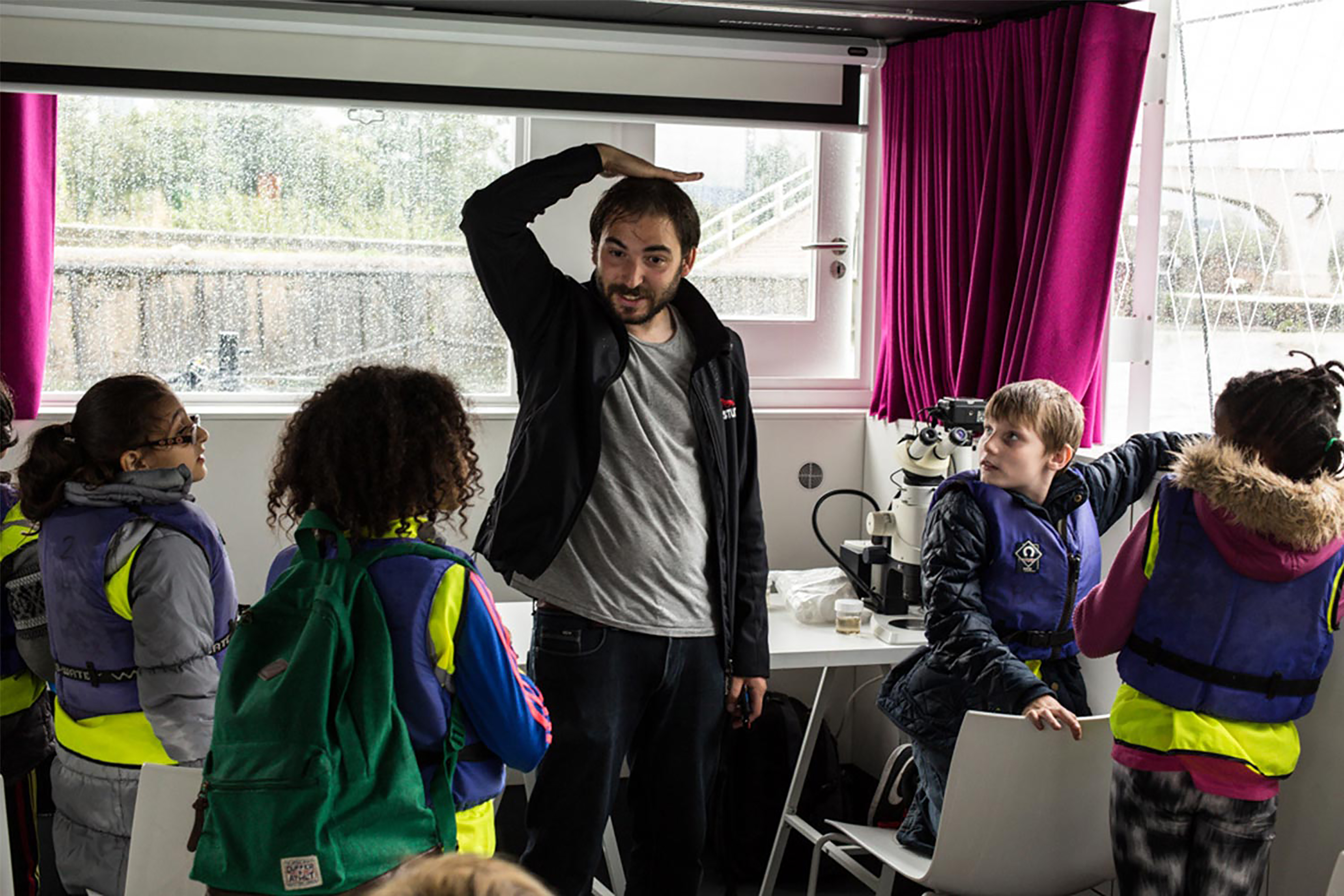 A man puts his right hand on his head while talking to a class of children wearing green Hi-Vis jackets indoors.