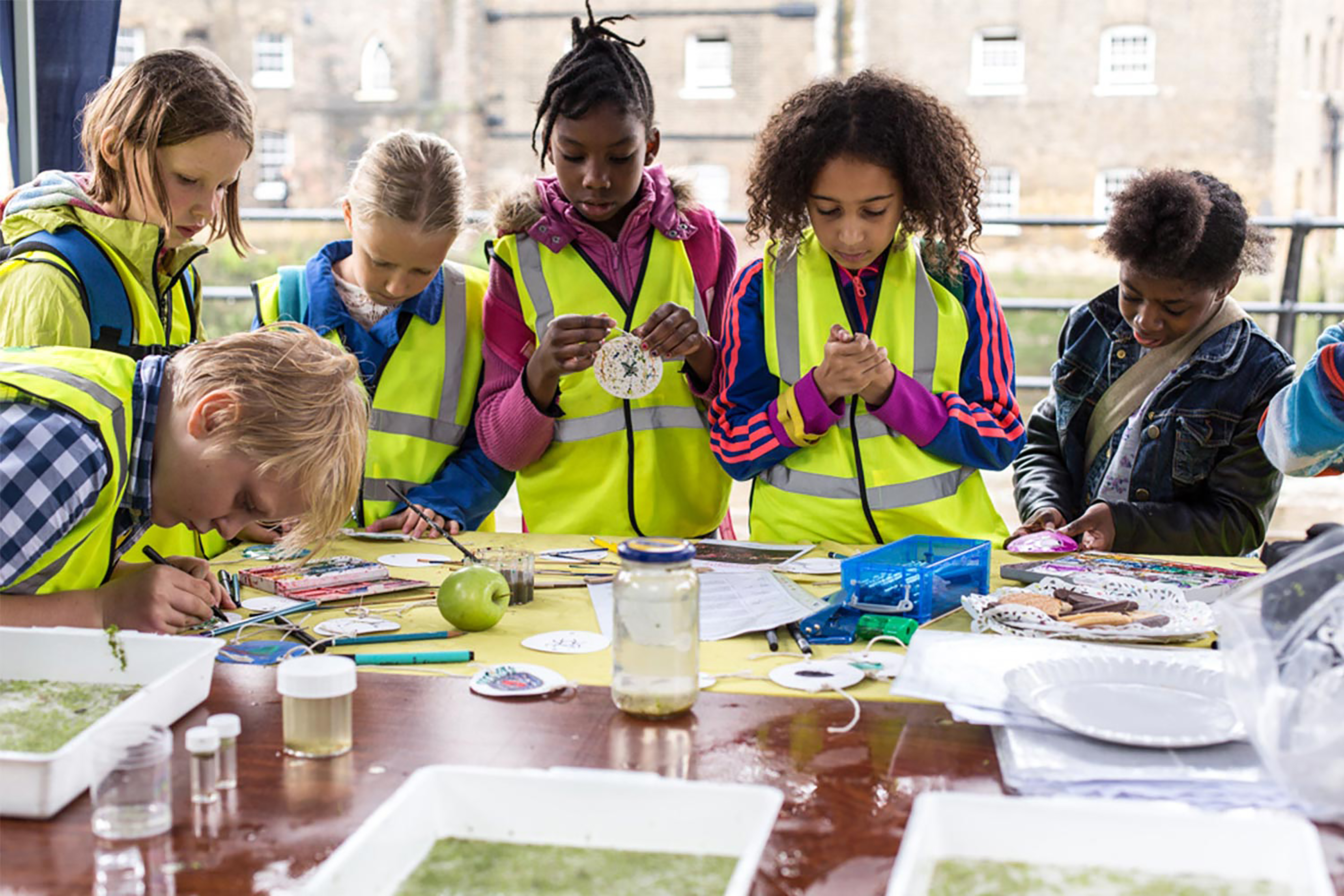Multiple children wearing green Hi-Vis jackets gathering round a table to handcraft items.