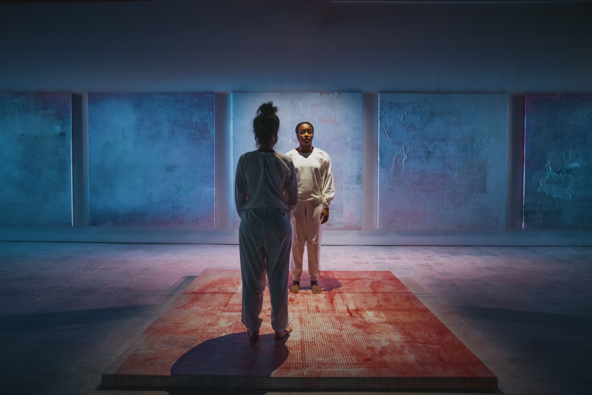Two women standing in a darkly lit room filled with a mix of blue, purple and pink colours, with both standing on a rusty pink textured flooring facing each other in loose baggy tops and sweatpants.