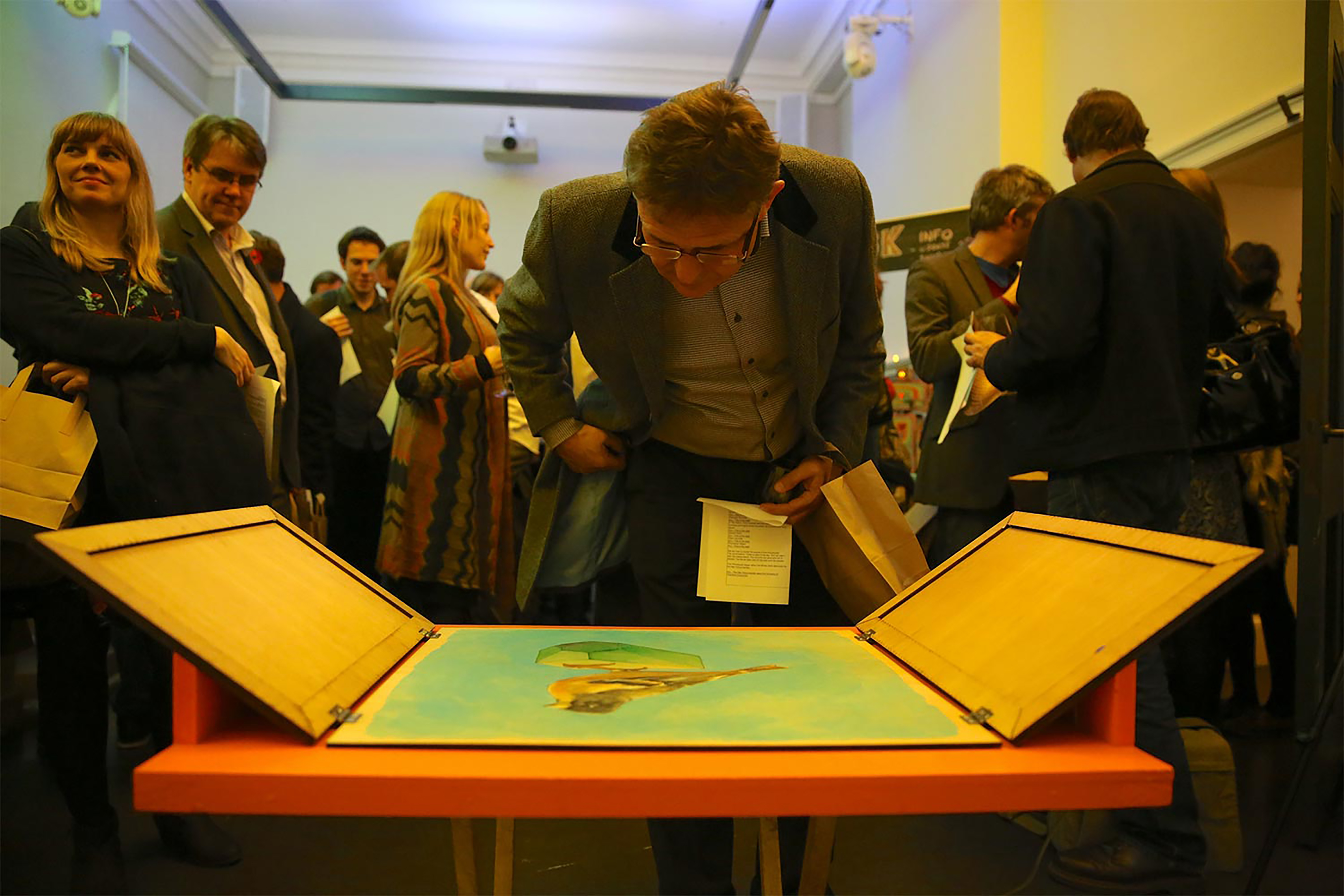 A middle age man wearing glasses and a suit jacket inspecting a canvas presented on flat plinth in front of a crowd of people talking.