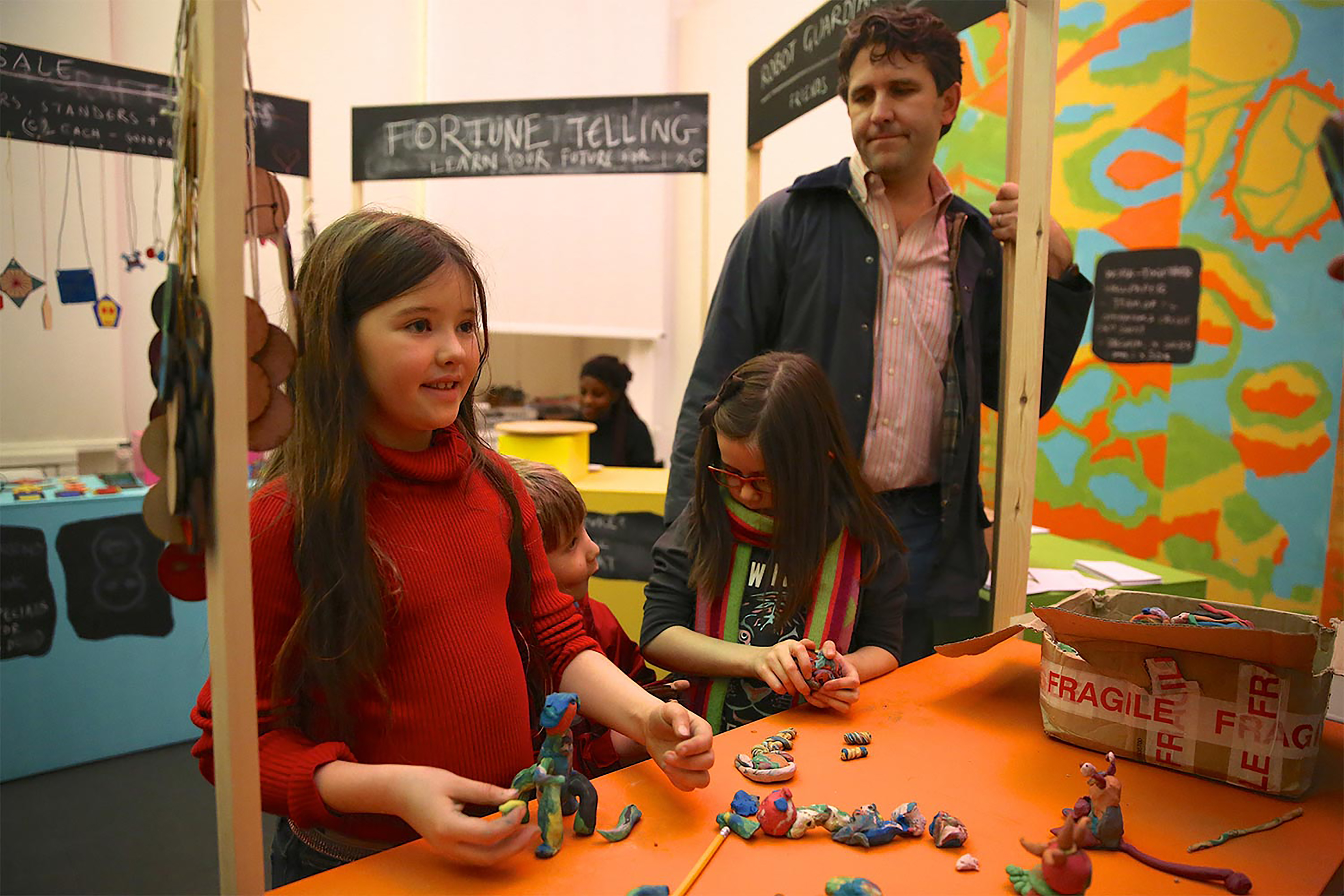 A photograph of children playing with plasticine models while a middle-aged man watches inside a classroom.