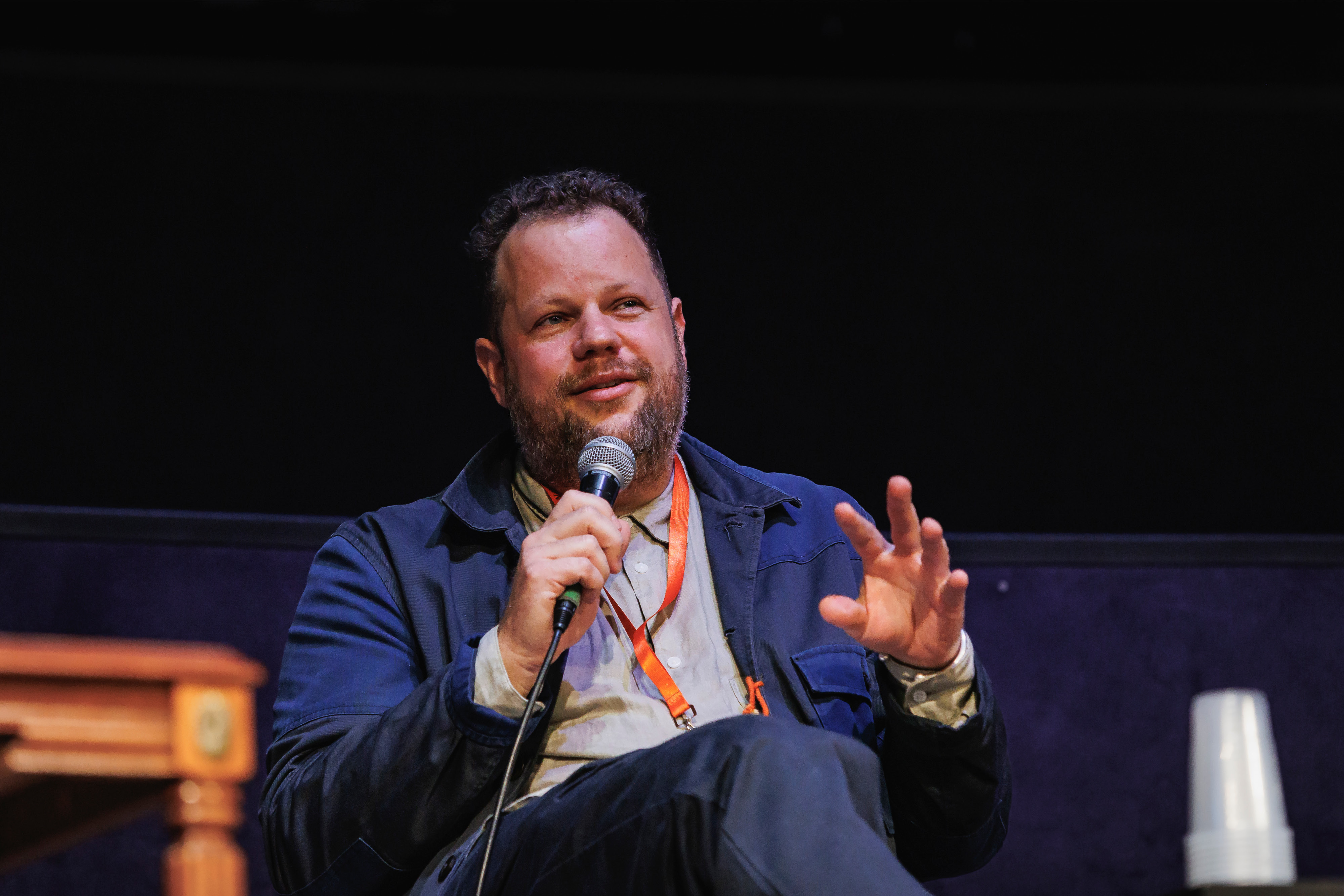 A man speaking on a stage holding a microphone. He is wearing a blue shirt, blue jacket and an orange lanyard.