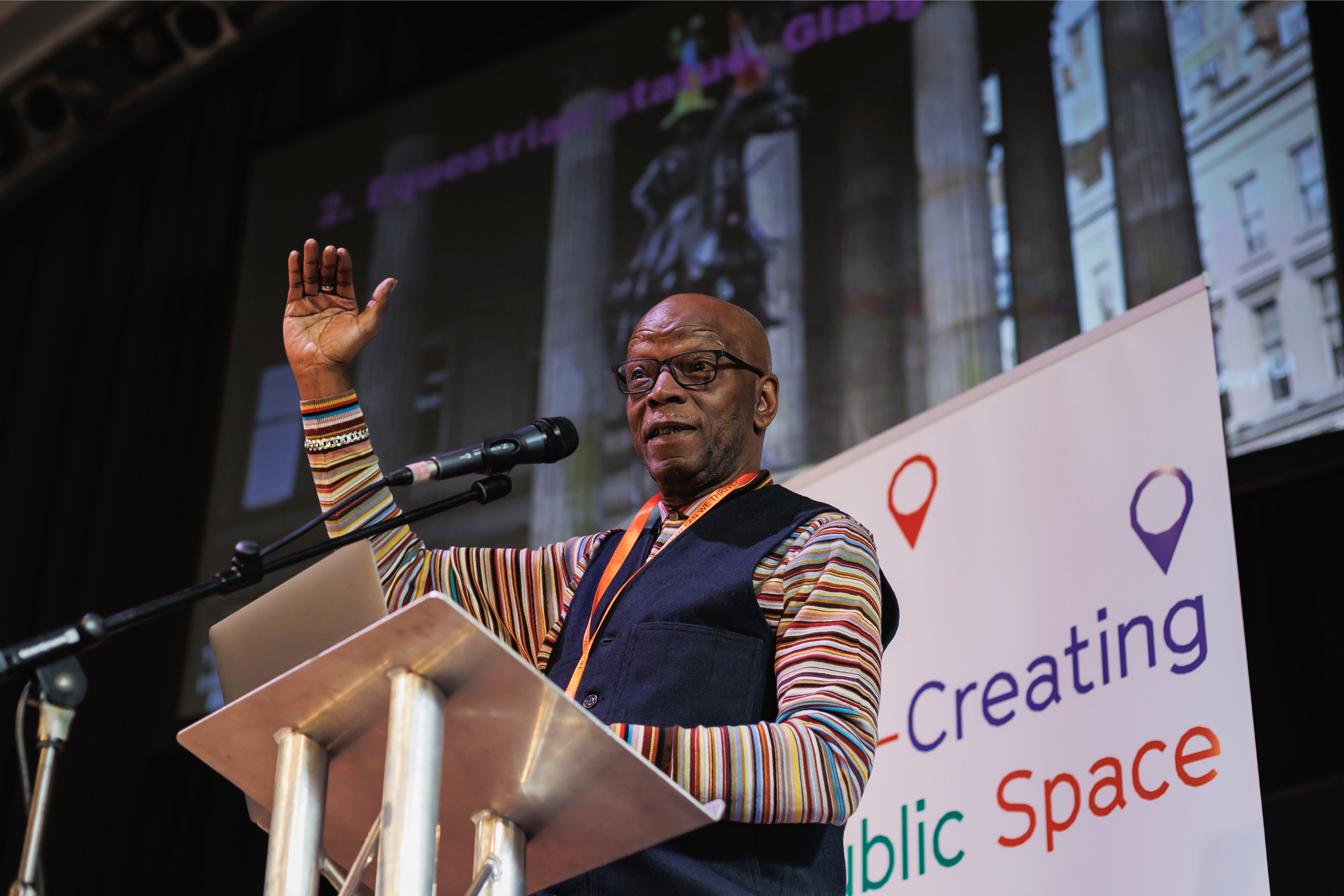 A bald black man speaking on a large stage behind a microphone stand. The stage has a large white banner, and a large screen behind them with an image of a grey building.