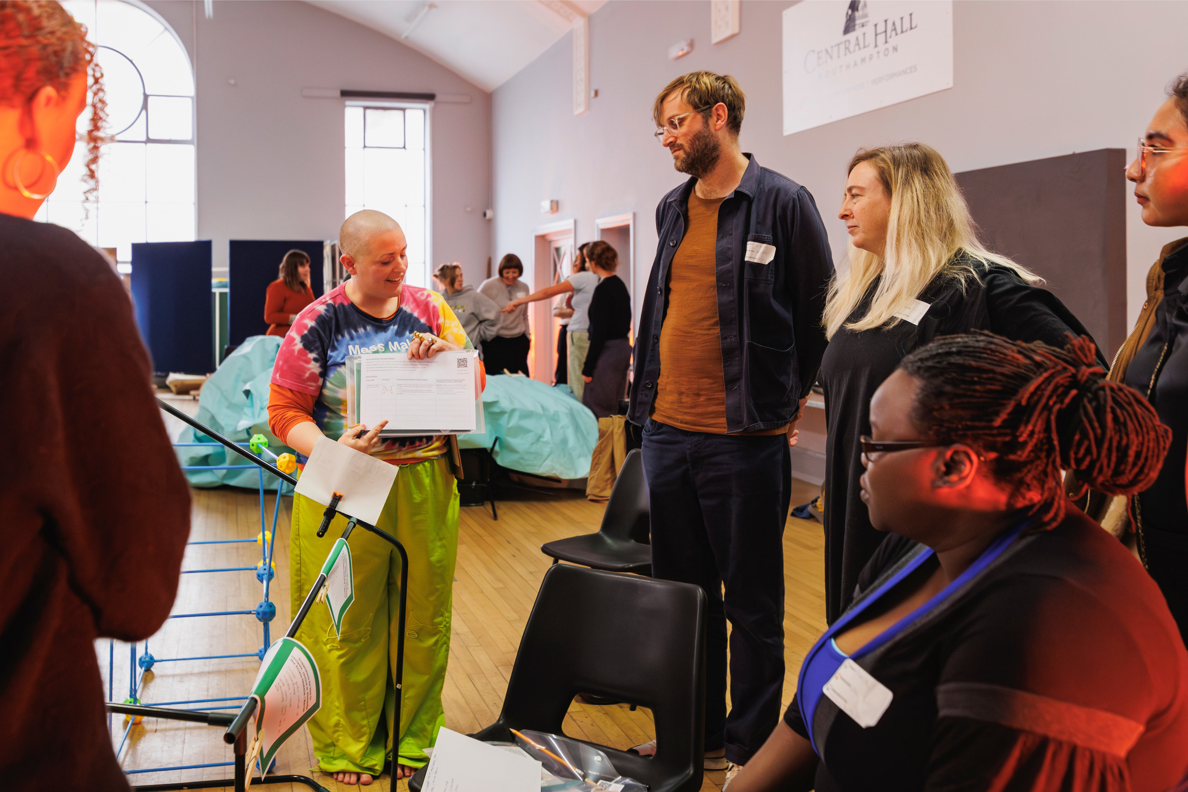 A group of people standing in a circle with white name badges on. There is a person in a colourful tie-dye top speaking and holding a folder.