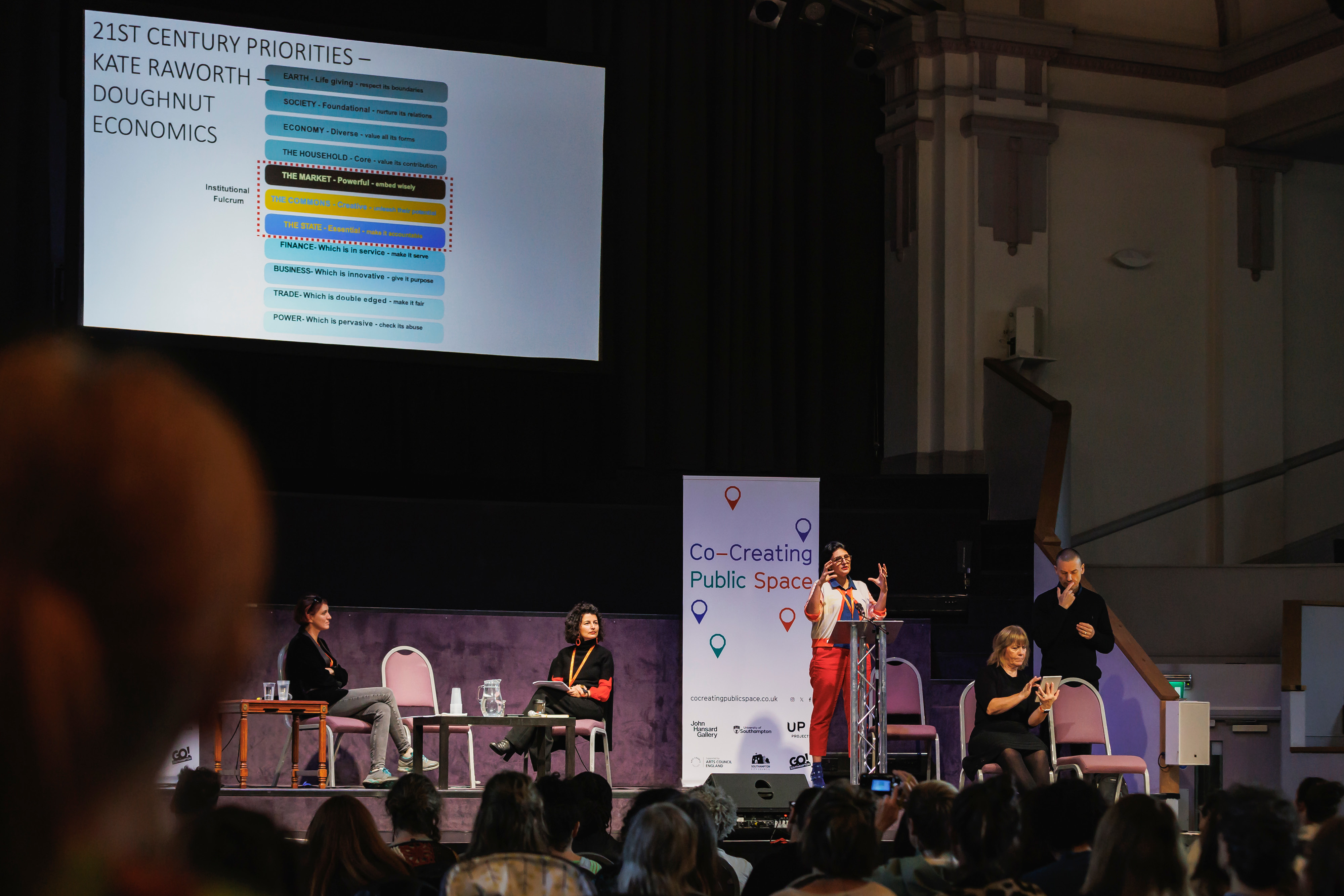 A crowd watching a woman presenting on a large stage behind a microphone lectern. The stage has a large white banner, various tables and chairs, and a large screen behind them with a graphic table of various words.