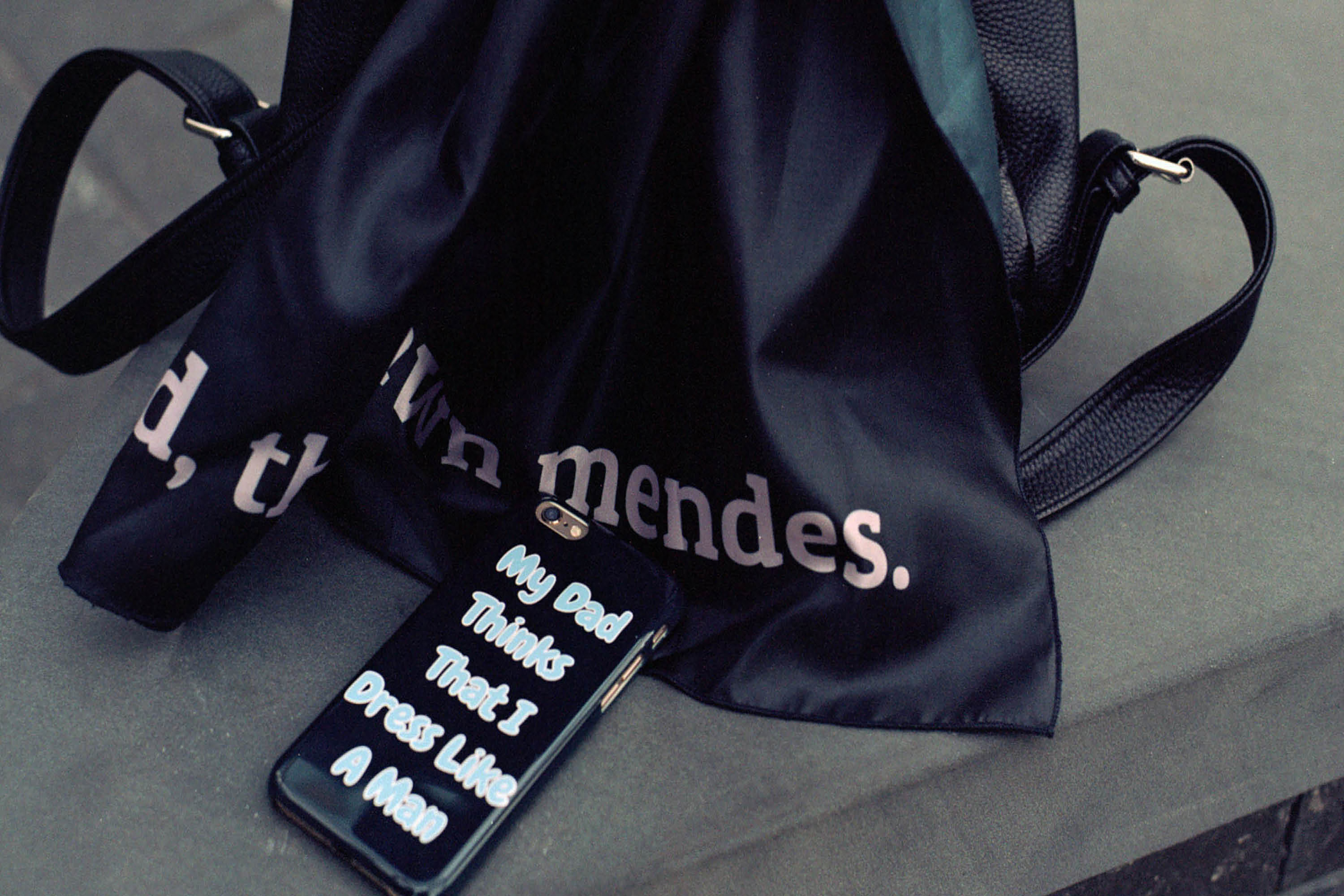 A black shoulder bag on the floor, with the words “Shawn Mendes”, next to a black phone case with the words “My dad thinks that I dress like a man”.