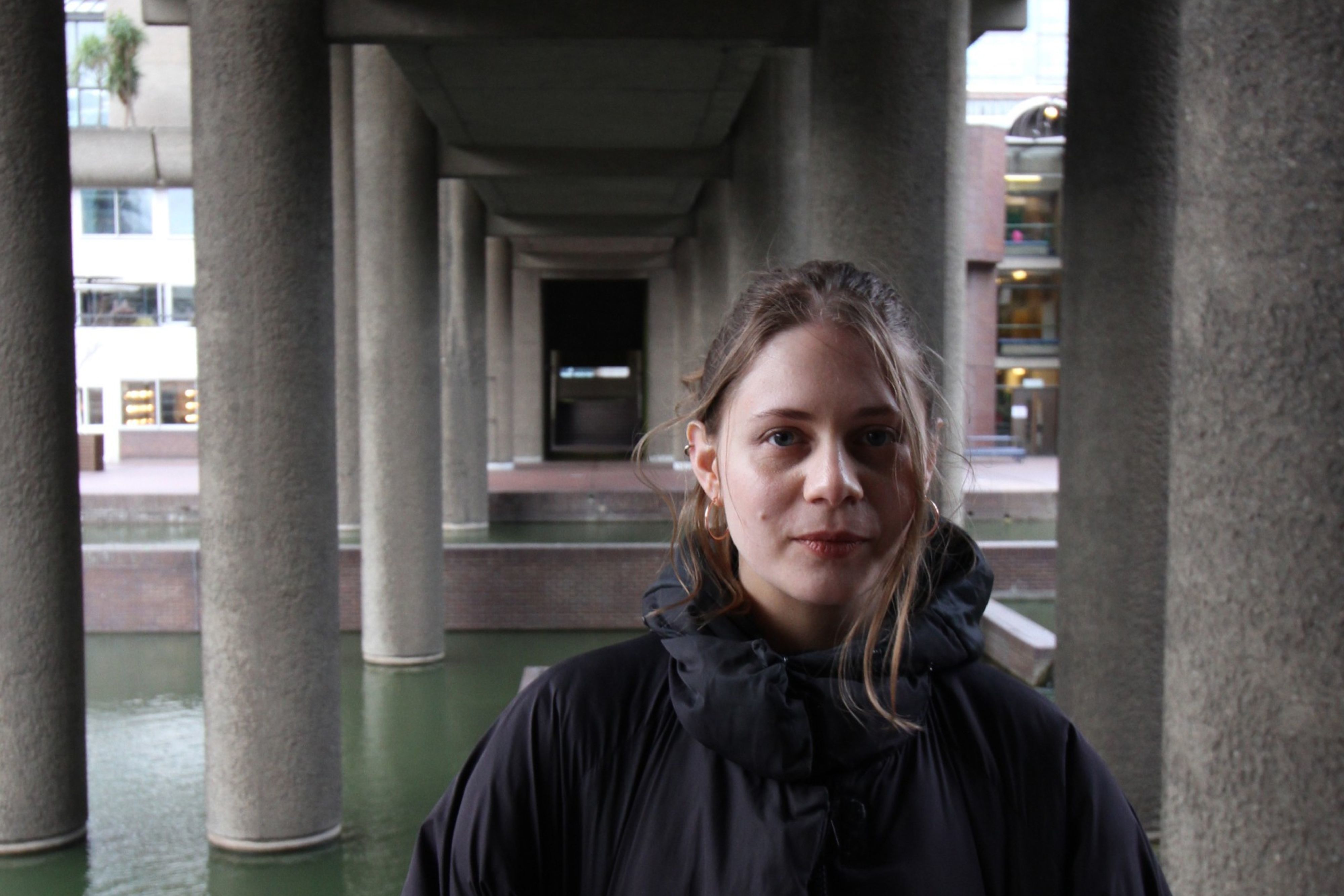Leah, a white woman in her 30s, smiles and looks at the camera. Her dark blonde hair is tied up but loose and windswept. She is wearing a large black coat and gold earrings, behind her are rows of concrete pillars receding into the background, holding up a roof over a green body of water.